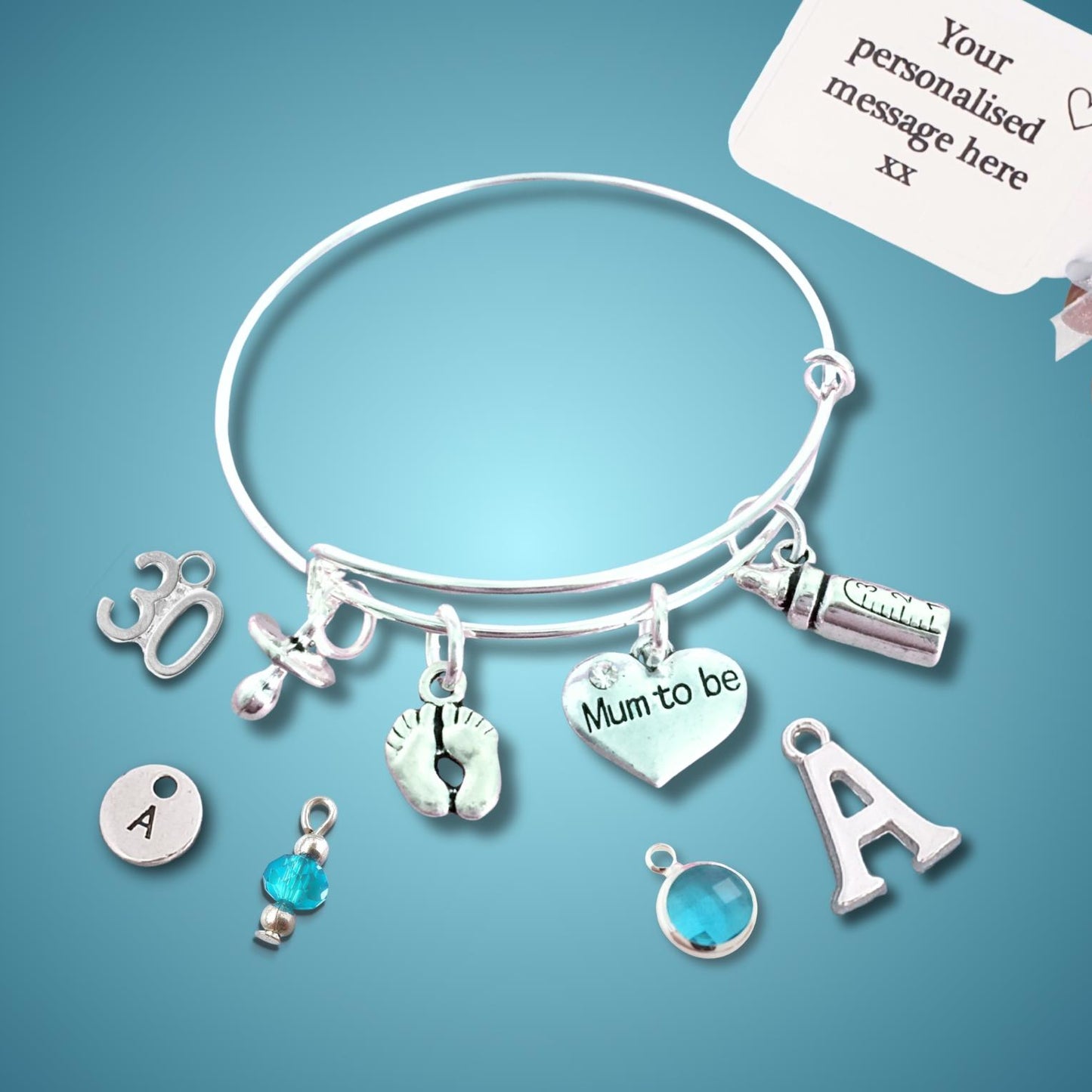 A silver charm bangle bracelet displayed on a blue gradient background, with a hand holding a white sample tag in the top right corner. The bracelet features multiple charms, including an initial 'A' disc, a baby footprint, a heart engraved with 'Mum to be', and other silver charms.