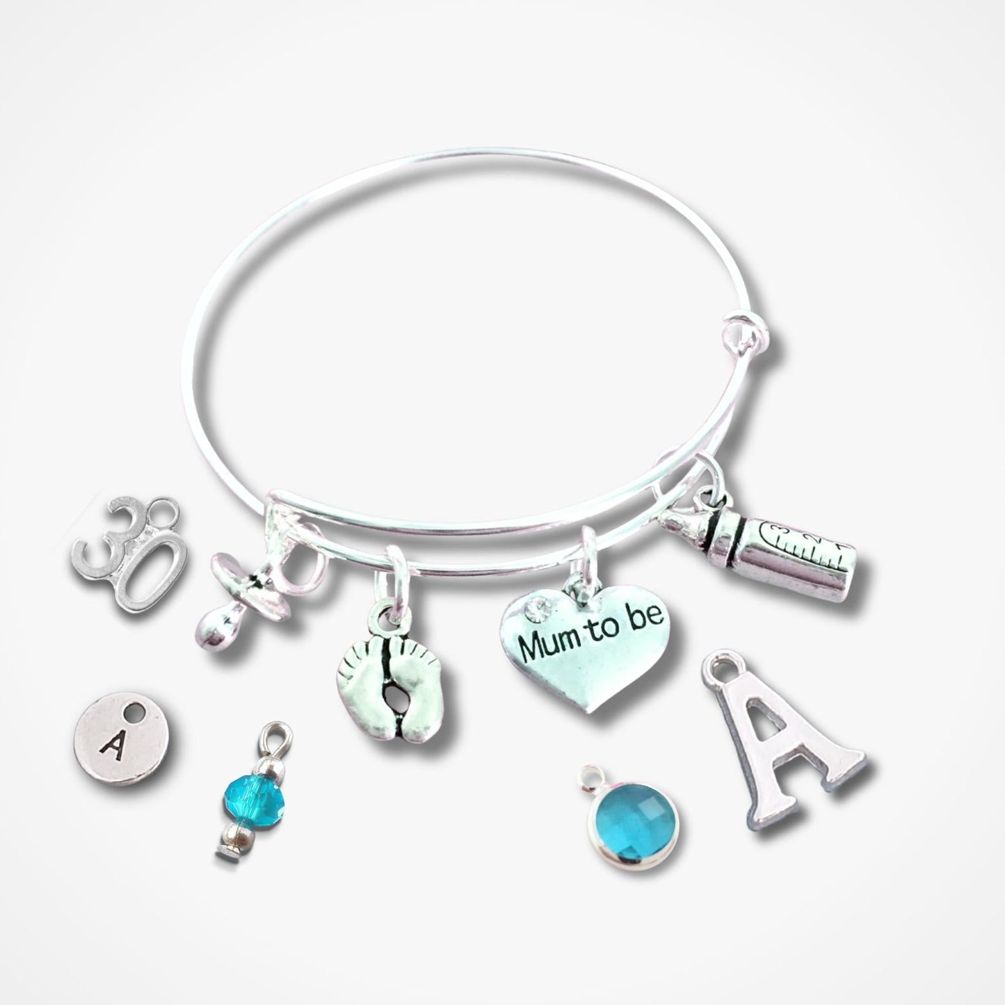 A silver bangle bracelet with multiple charms, including a heart charm that reads 'Mum to be', an initial 'A' charm, a small blue bead charm, and other silver-toned symbols, shown on a light gray background.