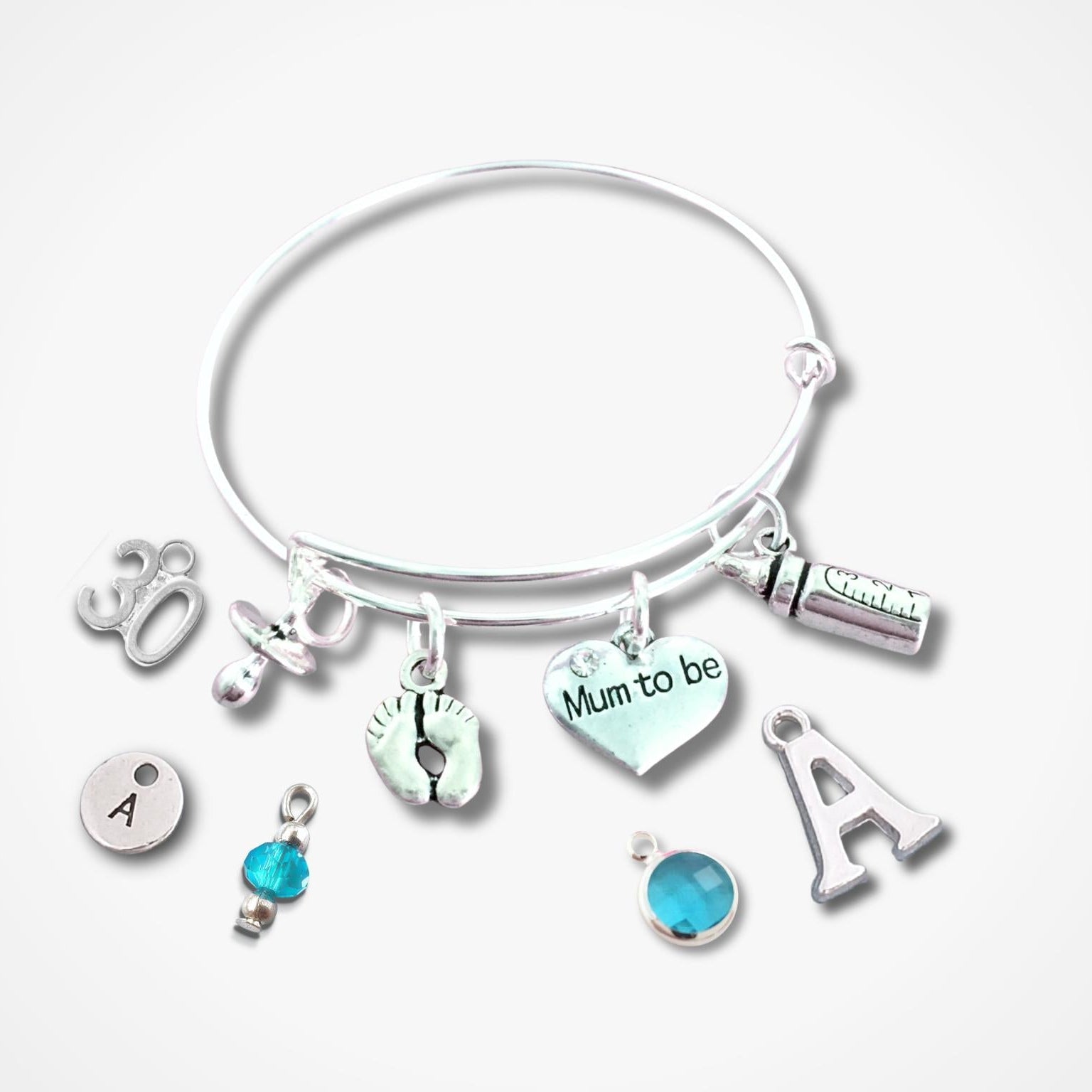 A silver bangle bracelet with multiple charms, including a heart charm that reads 'Mum to be', an initial 'A' charm, a small blue bead charm, and other silver-toned symbols, shown on a light gray background.