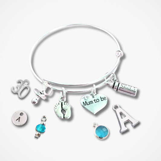 A silver bangle bracelet with multiple charms, including a heart charm that reads 'Mum to be', an initial 'A' charm, a small blue bead charm, and other silver-toned symbols, shown on a light gray background.