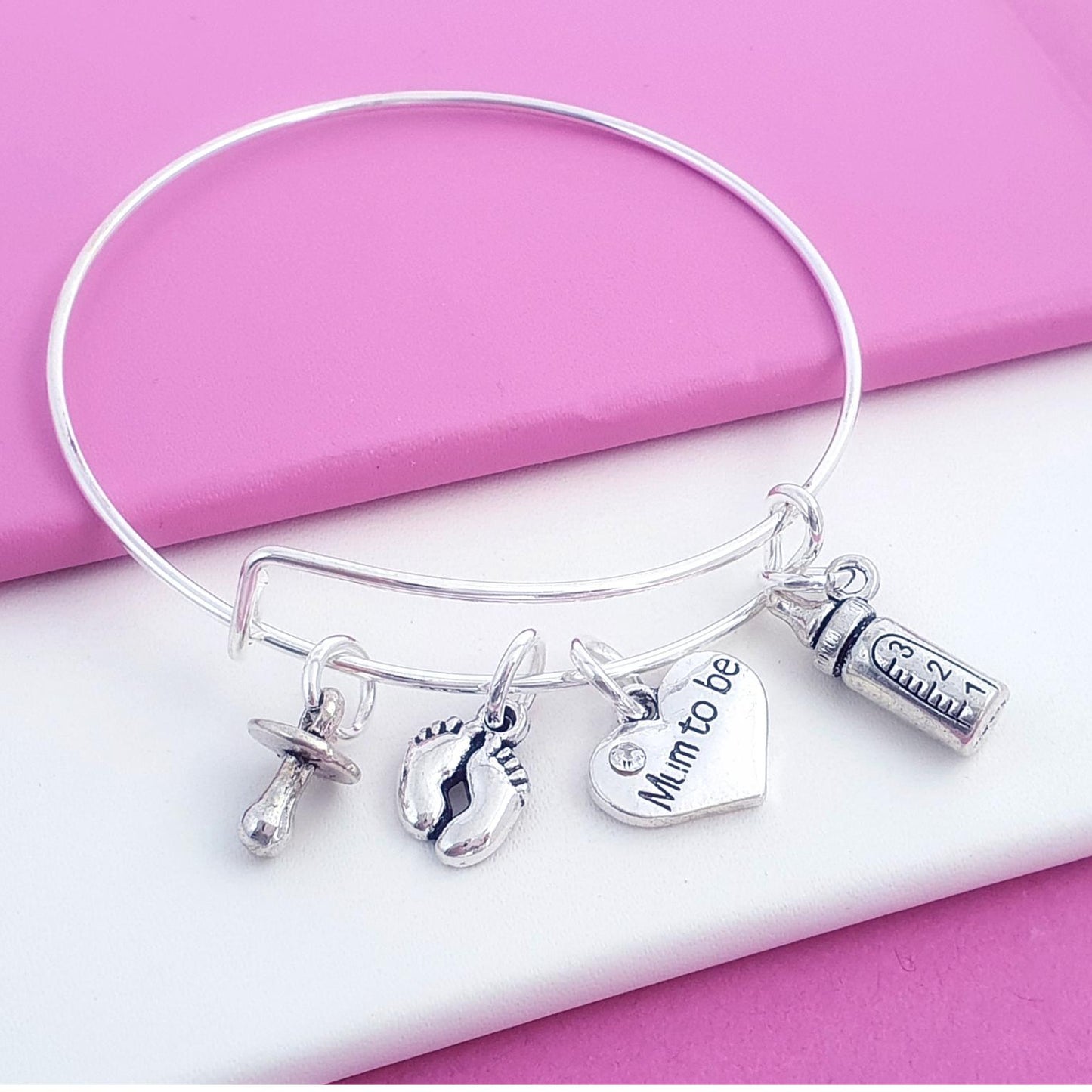 A silver-tone bangle bracelet with four charms, including a heart charm engraved 'Mum to be', a baby bottle charm, and a pacifier charm, displayed on a pink and white background.