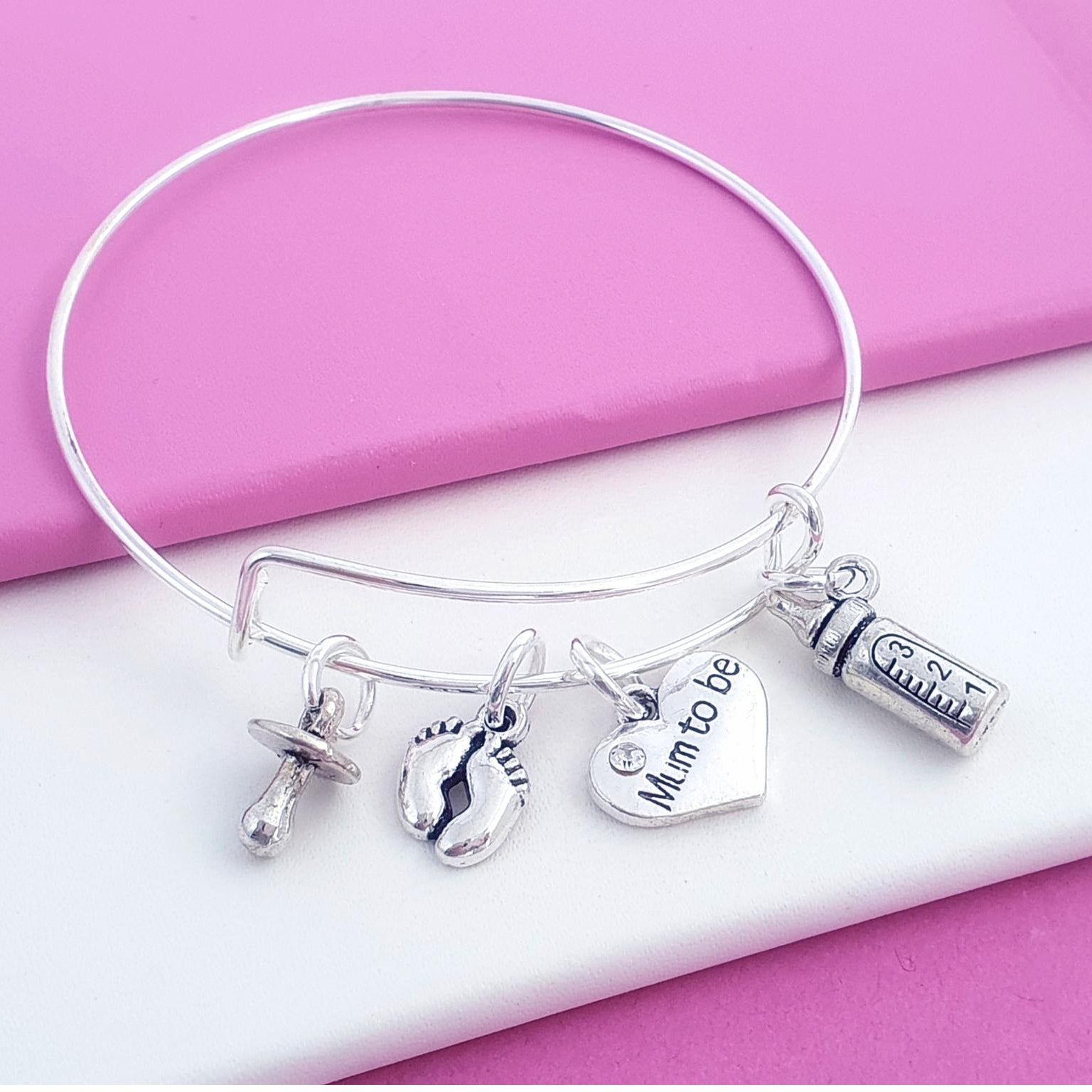 A silver-tone bangle bracelet with four charms, including a heart charm engraved 'Mum to be', a baby bottle charm, and a pacifier charm, displayed on a pink and white background.