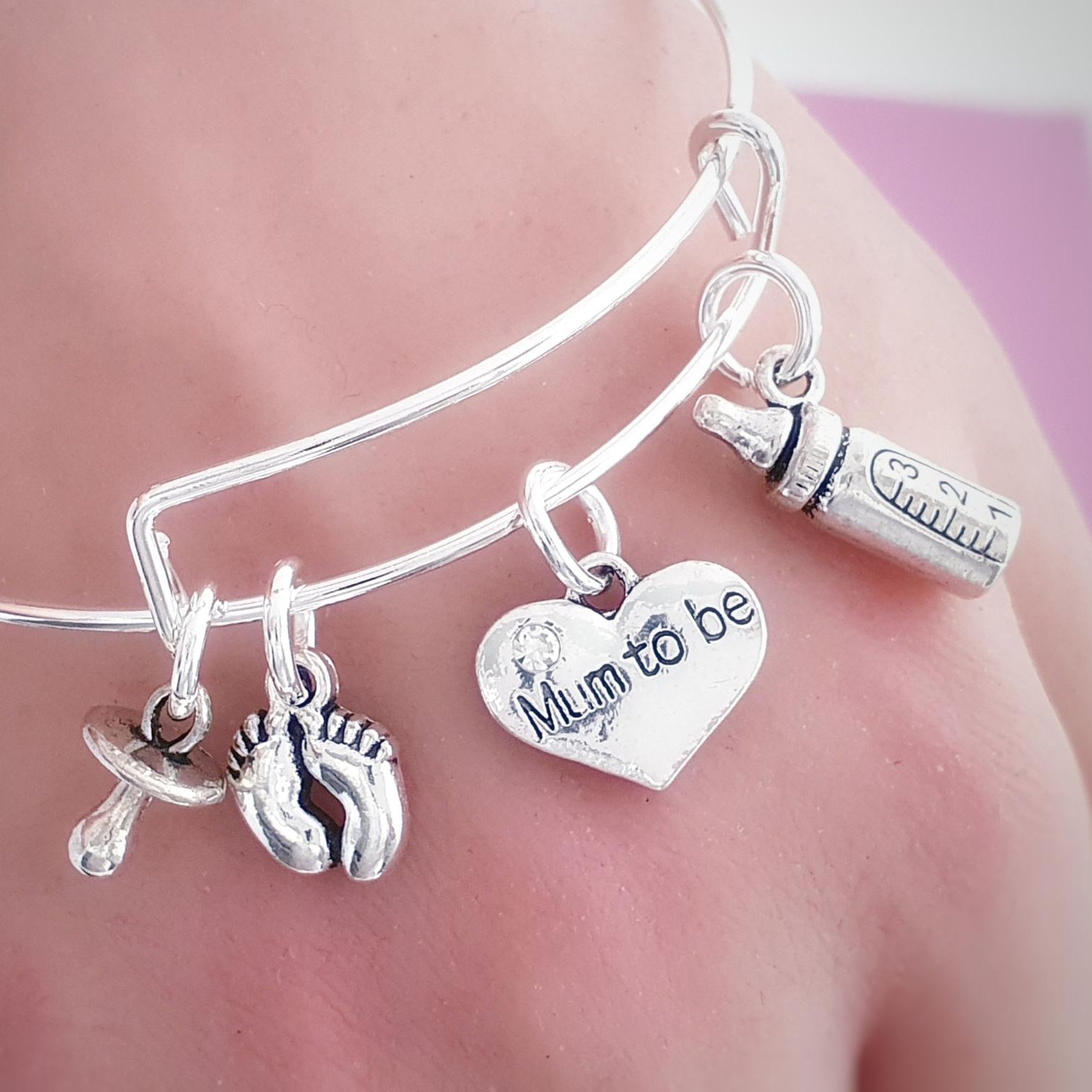 A close-up of a silver bangle bracelet worn on a wrist, featuring charms including baby footprints, a heart engraved with 'Mum to be', a pacifier, and a baby bottle.
