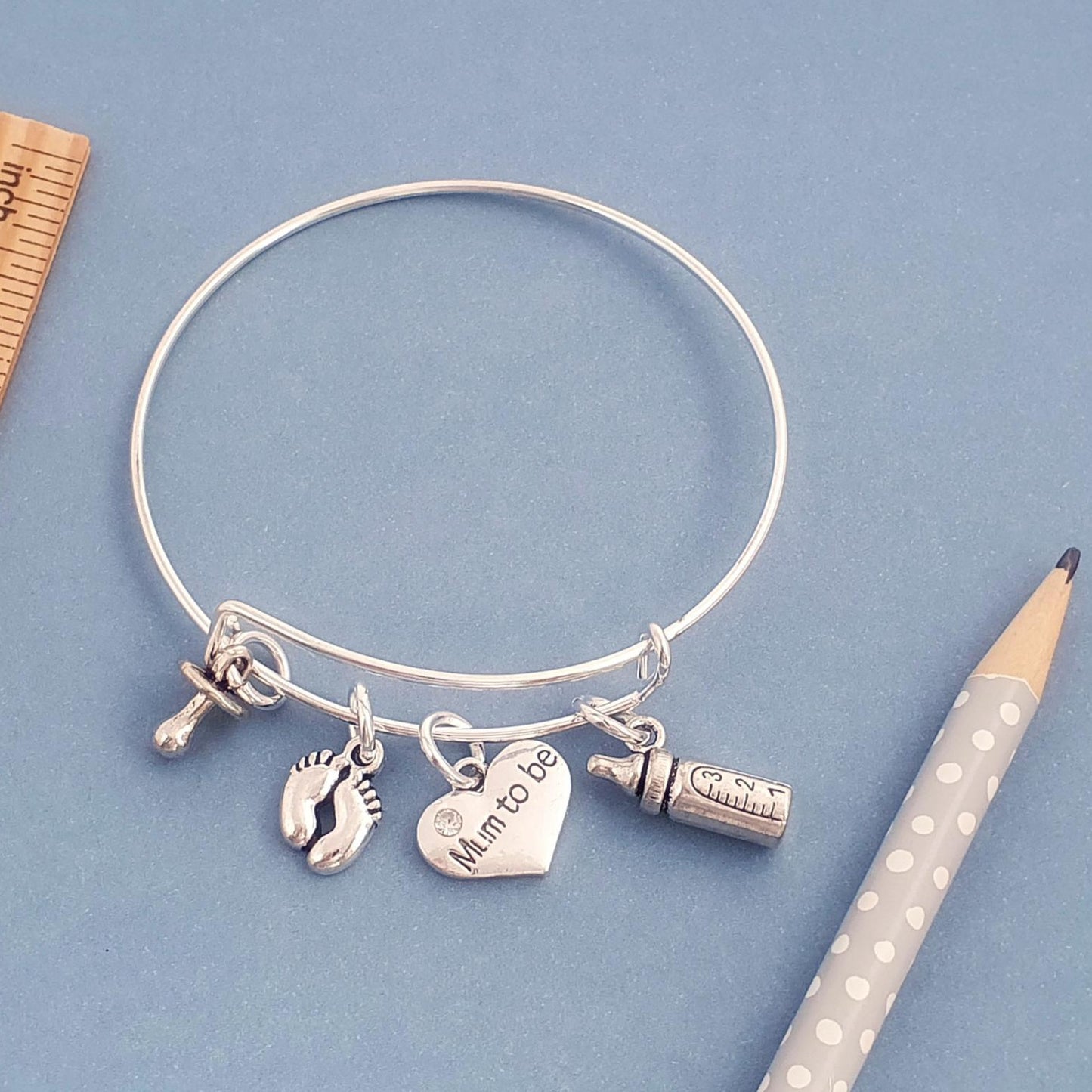 A silver bangle bracelet with charms including a heart that says 'Mom to be', a baby foot, and a bottle, displayed on a blue background with a wooden ruler and a pencil.