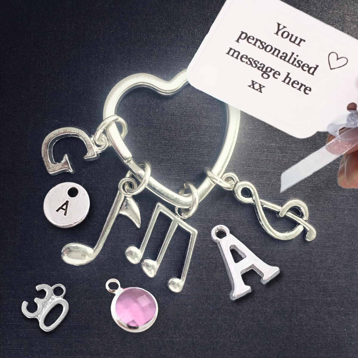A silver-tone personalized charm keyring with musical notes, letters, and decorative symbols, shown on a dark background with a white sample tag and a hand holding the tag.