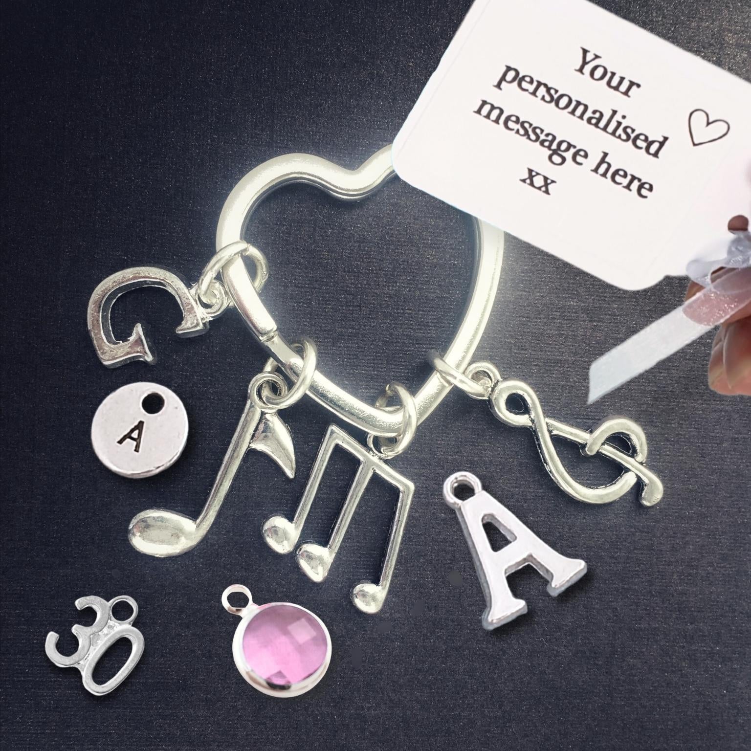 A silver-tone personalized charm keyring with musical notes, letters, and decorative symbols, shown on a dark background with a white sample tag and a hand holding the tag.