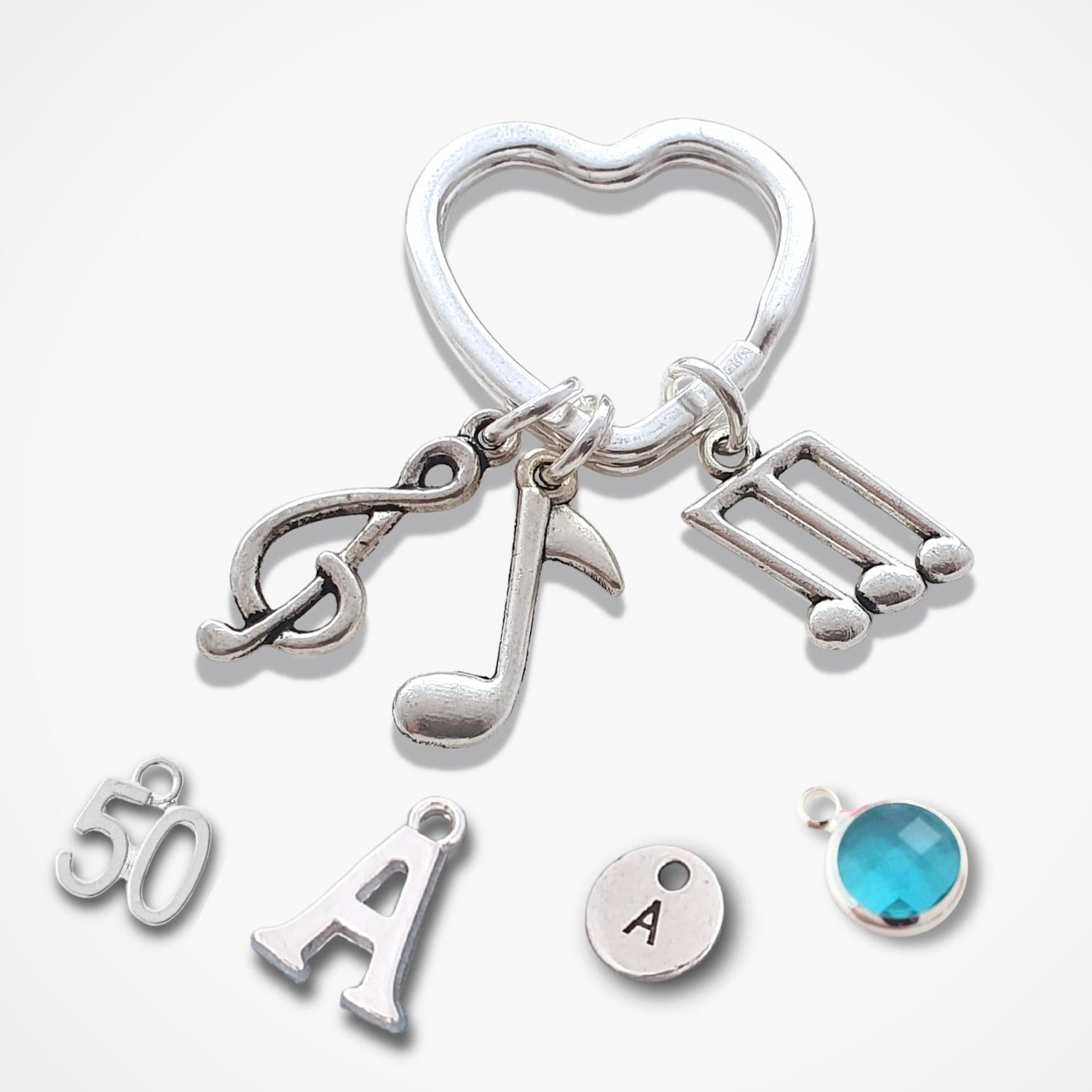 A silver-toned jewelry keyring charm set featuring a heart-shaped ring, music note and treble clef charms, a double-strike musical note charm, a '50' charm, an engraved letter 'A' charm, a round silver disc charm with the letter 'A', and a small round turquoise-blue faceted bead charm, all arranged on a light gray background.
