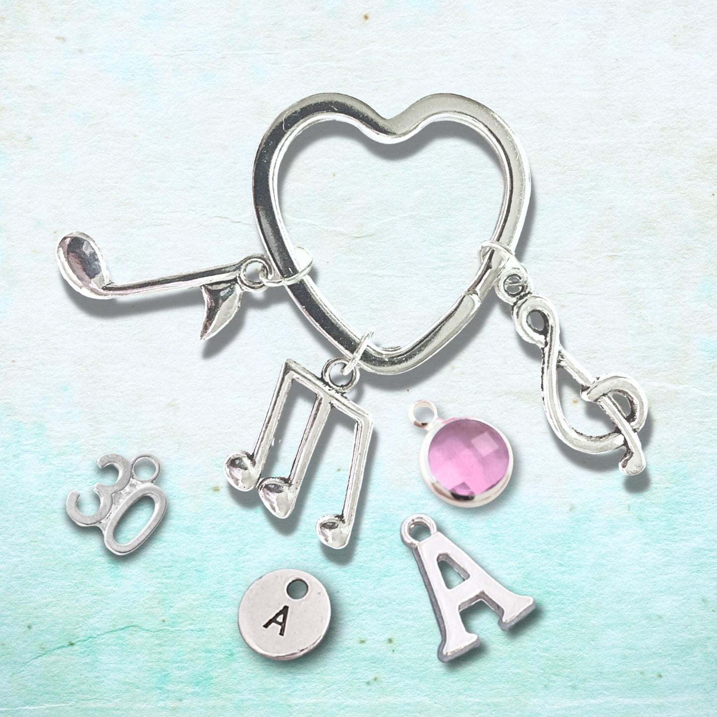 A silver-toned jewelry charm keyring featuring a heart-shaped clasp and dangling charms including musical notes, treble and bass clefs, a pink oval gemstone, a round silver disc stamped with 'A', and a letter 'A' charm, displayed on a light blue textured background.