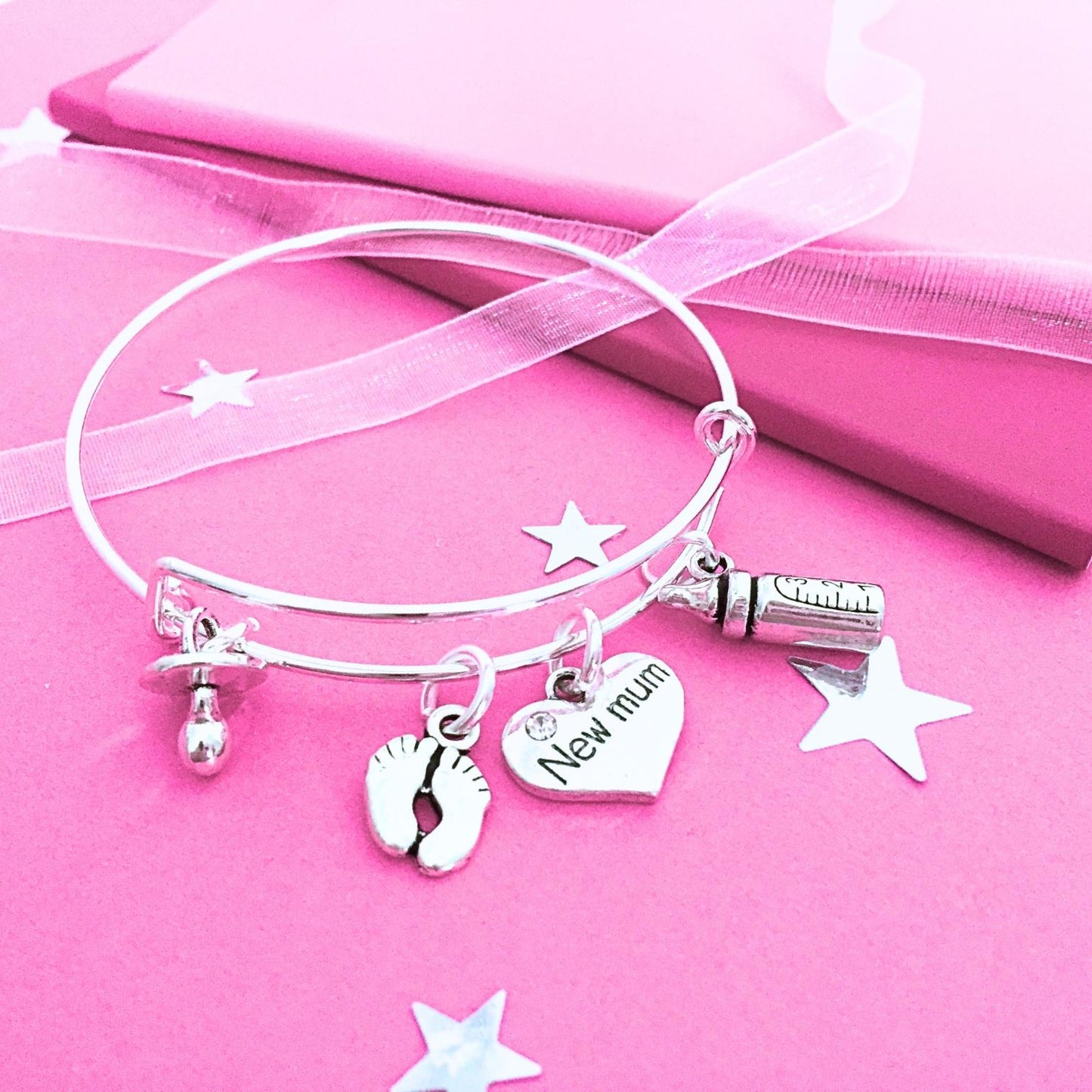 A silver bangle bracelet with charms, including a heart charm engraved 'New mum', a baby bottle charm, and a footprints charm, displayed on a pink background with star cutouts and a pink gift box.
