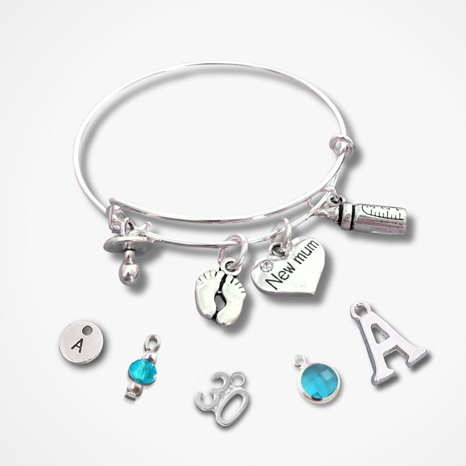 A silver bangle bracelet with multiple charms, including a heart charm engraved 'New mum', a footprint charm, a baby bottle charm, and several letter and gemstone charms, displayed on a light gray background.