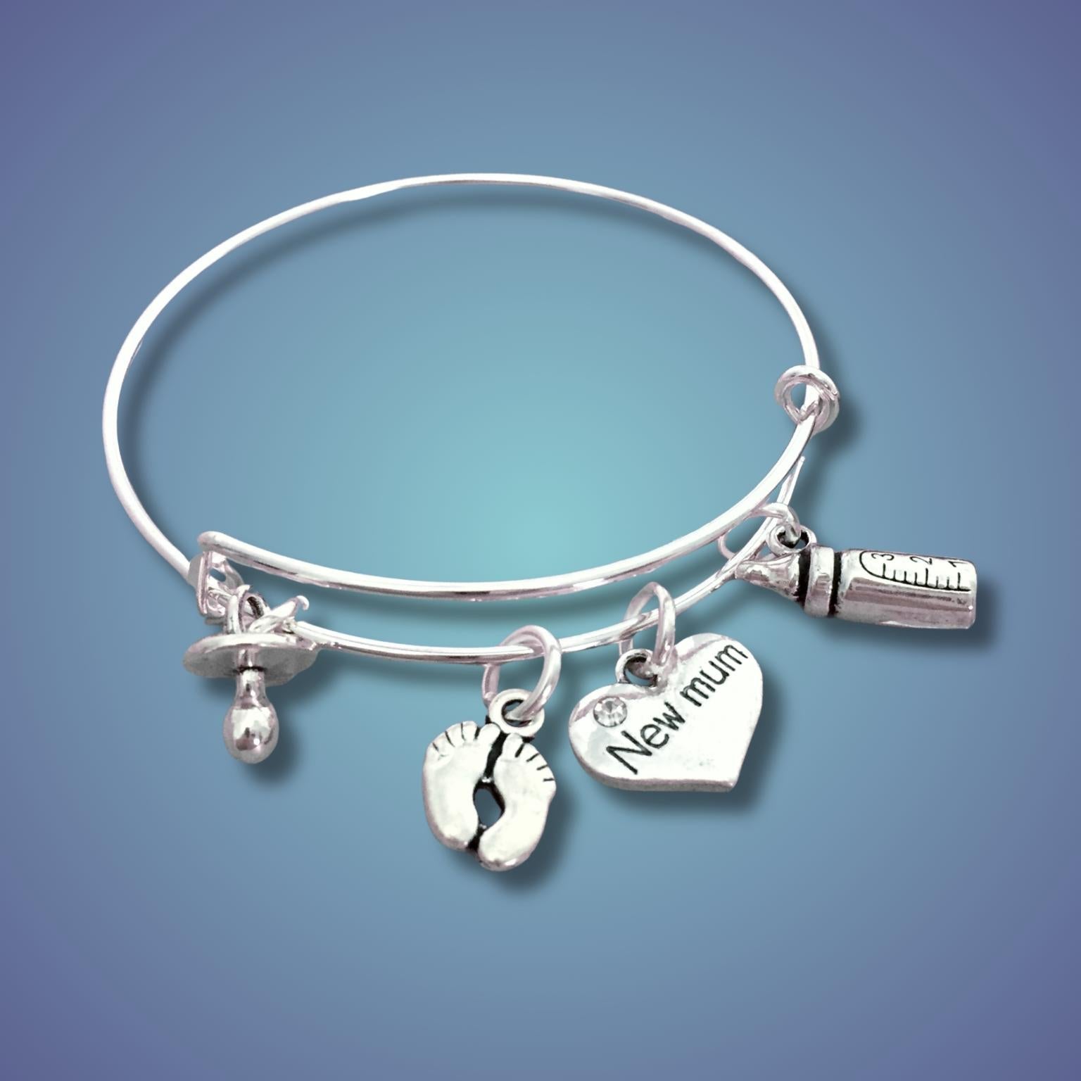 A silver bangle bracelet with three dangling charms: a baby bottle, a footprint, and a heart engraved with 'New mum', shown on a blue gradient background.