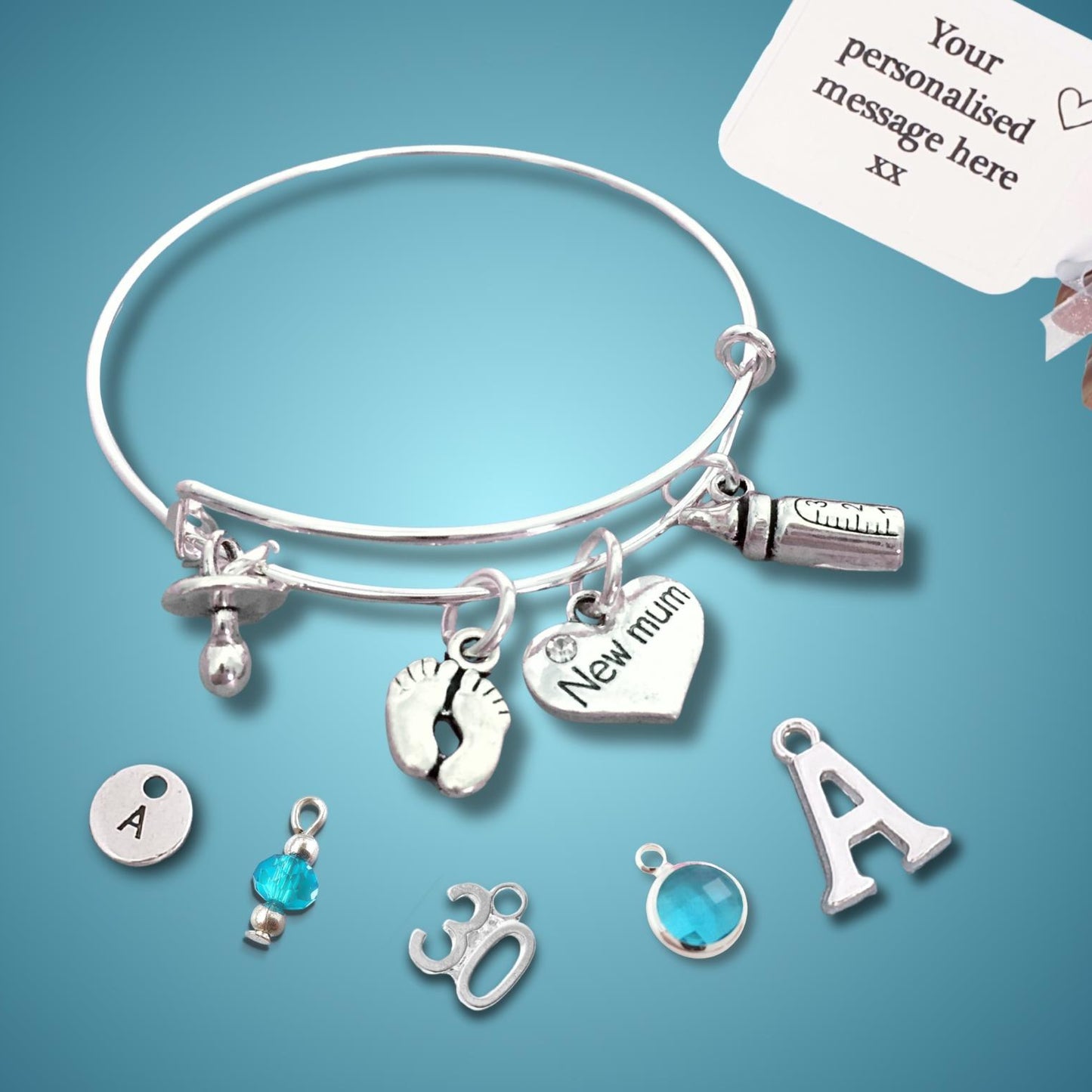A silver charm bracelet displayed on a blue gradient background, with a hand holding a white personalization card in the top right corner. The bracelet features multiple charms including a heart engraved 'New mum', baby footprints, a bottle-shaped charm, and a capsule charm.