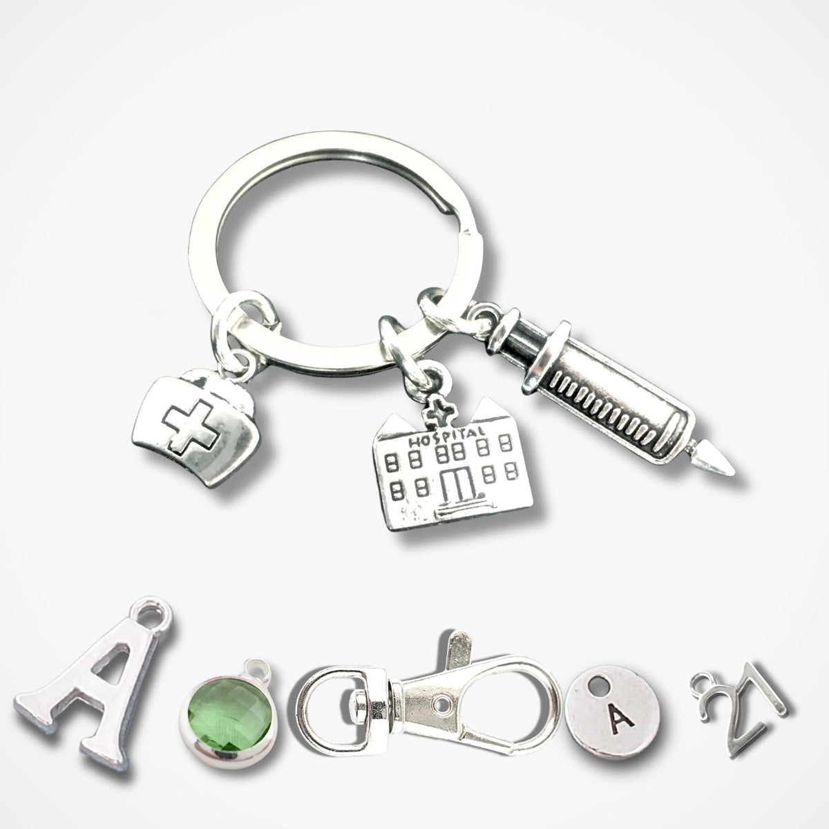A silver keychain set featuring medical-themed charms including a syringe, a hospital building, and a nurse hat, shown alongside individual letter 'A' charms, a green gemstone, and a clasp on a light gray background.