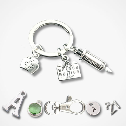 A silver keychain set featuring medical-themed charms including a syringe, a hospital building, and a nurse hat, shown alongside individual letter 'A' charms, a green gemstone, and a clasp on a light gray background.