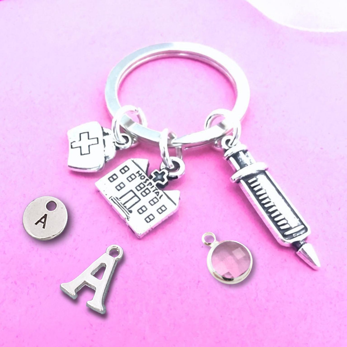 A silver keychain with medical-themed charms, including a syringe, hospital, nurse bag, and letter 'A', displayed on a pink background.