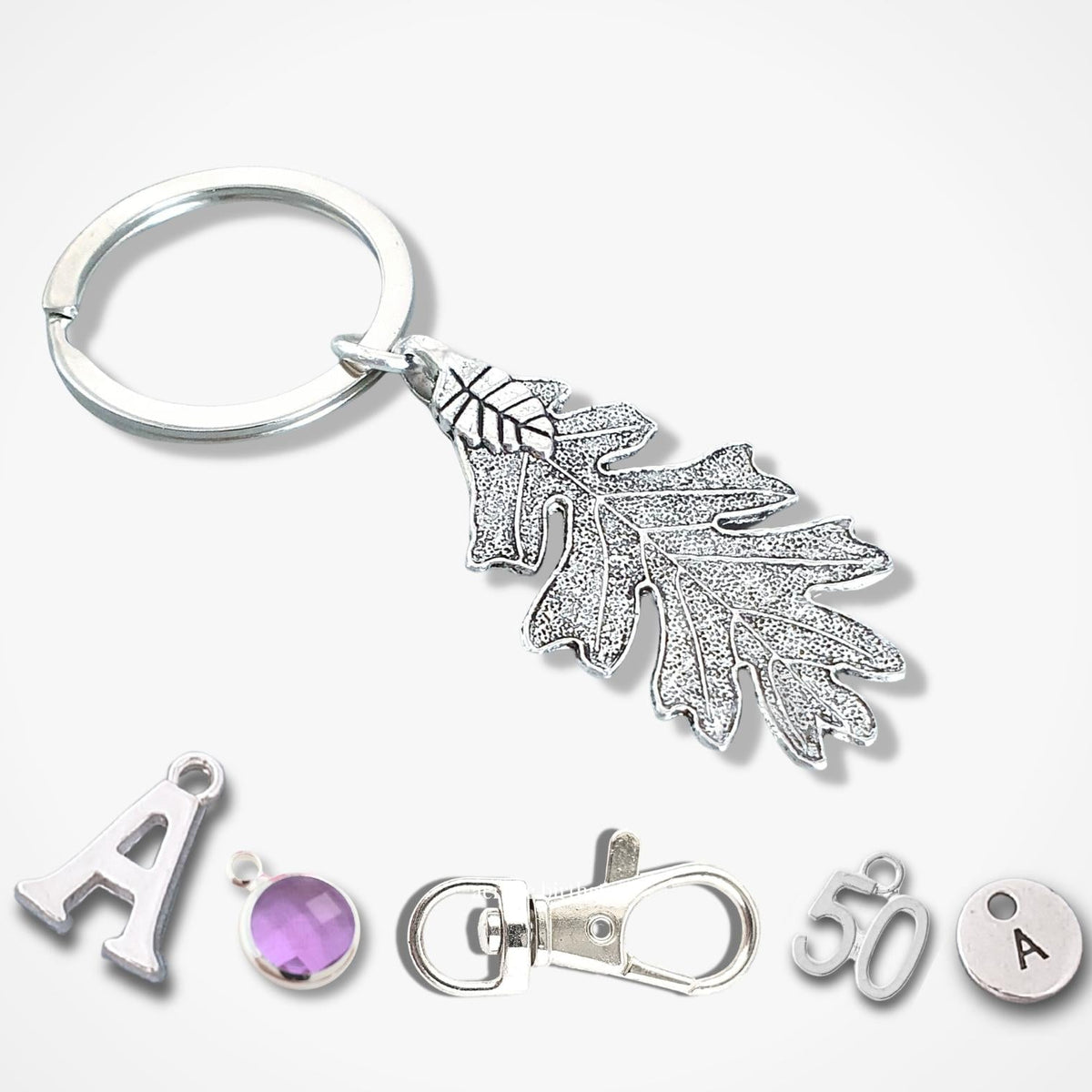 Silver metal keychain set featuring a leaf-shaped charm with a textured, finish, a split key ring, a lobster clasp, and small letter 'A' charms with a purple gemstone and a round silver disc, all arranged on a light gray background.
