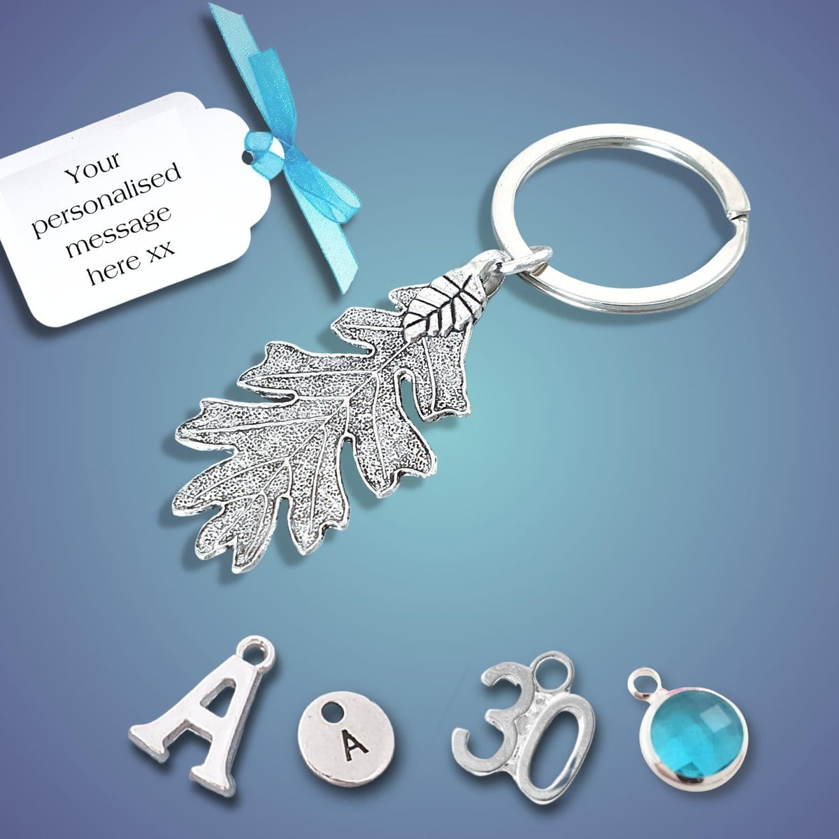 A silver oak leaf keychain with a split ring, shown on a blue gradient background with a white tag that reads 'Your personalised message here xx' and four small charm pieces below: a letter A, a round letter A disc, a number 30, and a round blue gemstone.