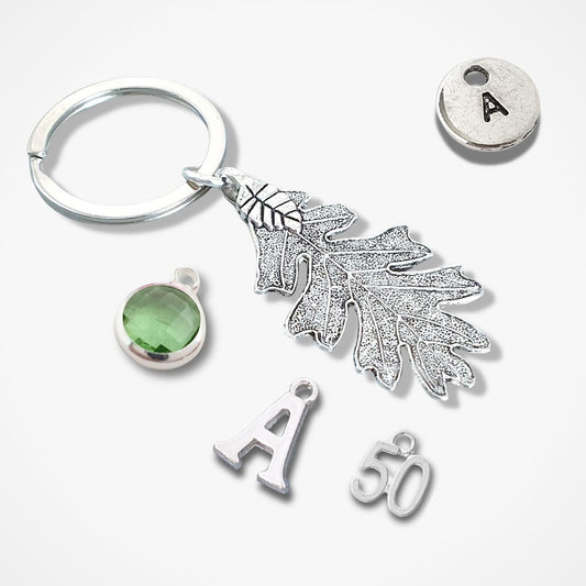 A silver keychain set featuring an oak leaf charm, a split key ring, a round silver disc engraved with the letter 'A', a green oval stone charm, and two letter 'A' charms, all arranged on a light gray background.