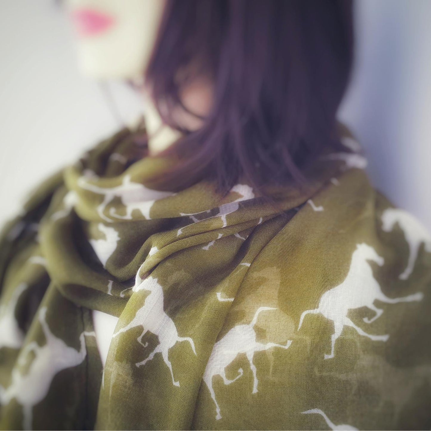 An olive green scarf with a white horse pattern, worn by a model with dark hair in a close-up shot.