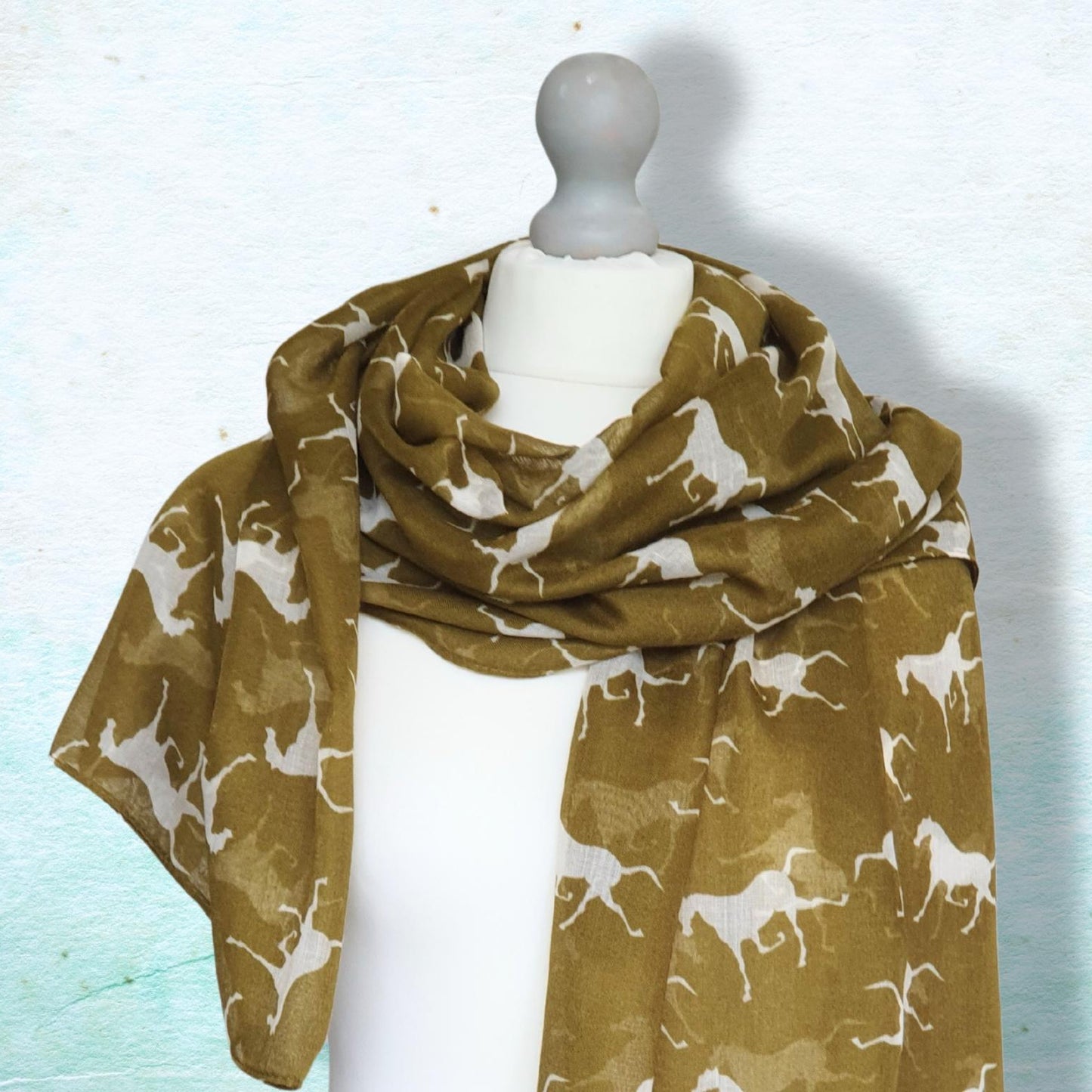 A lightweight scarf draped on a white mannequin torso, featuring an all-over print of white horses on an olive green background, set against a textured light blue wall.