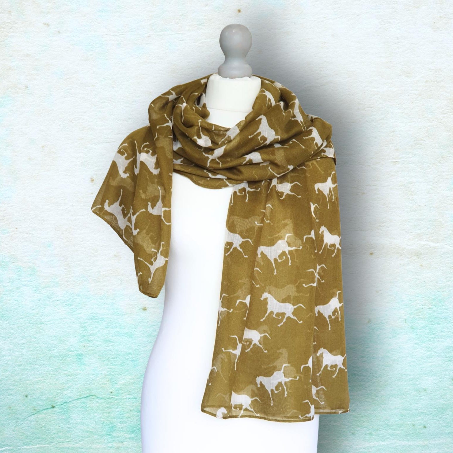 A lightweight scarf in a warm mustard-brown color with a repeating white pattern of running horses, displayed on a white mannequin torso against a textured light blue background.