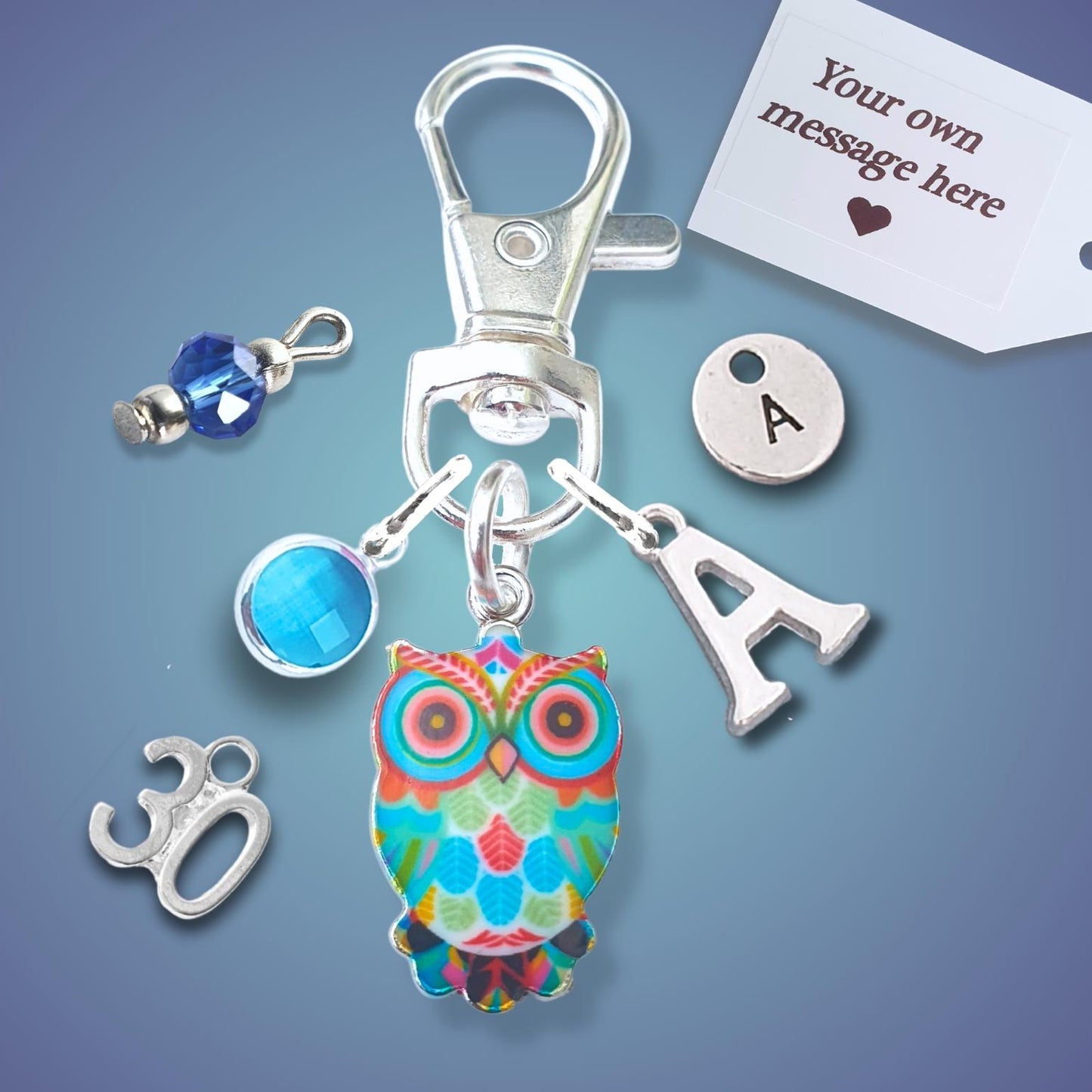 A silver-toned keychain or bag charm set on a blue gradient background, featuring a colorful owl charm, a silver letter 'A' charm, a blue faceted bead, a silver '30' charm, and a white customizable tag.