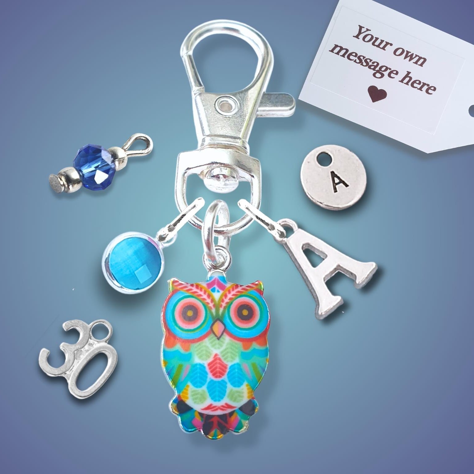 A silver-toned keychain or bag charm set on a blue gradient background, featuring a colorful owl charm, a silver letter 'A' charm, a blue faceted bead, a silver '30' charm, and a white customizable tag.