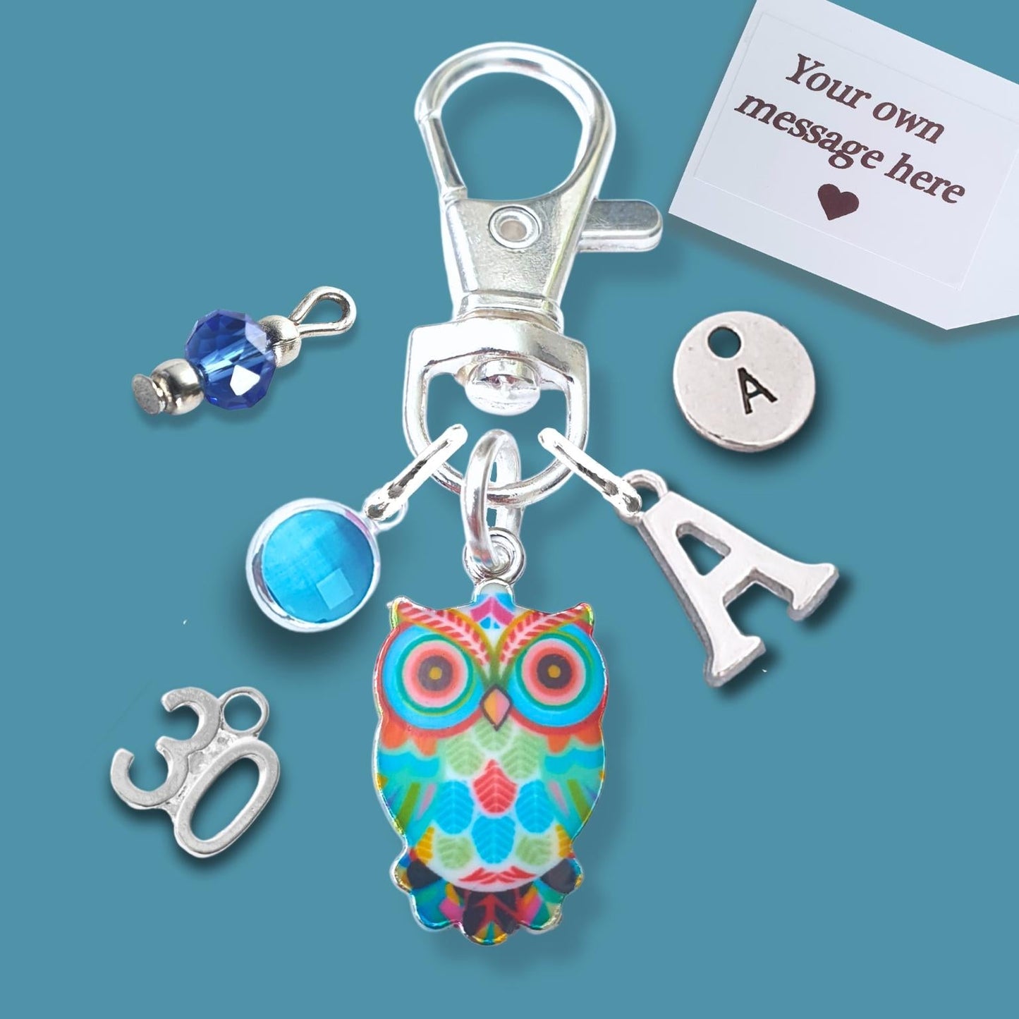 A silver lobster clasp keychain charm set on a teal background, featuring a colorful owl charm, a silver letter 'A' charm, a blue faceted bead, a silver '30' charm, and a white customizable message tag.
