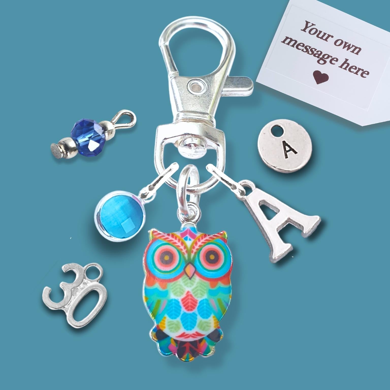 A silver lobster clasp keychain charm set on a teal background, featuring a colorful owl charm, a silver letter 'A' charm, a blue faceted bead, a silver '30' charm, and a white customizable message tag.