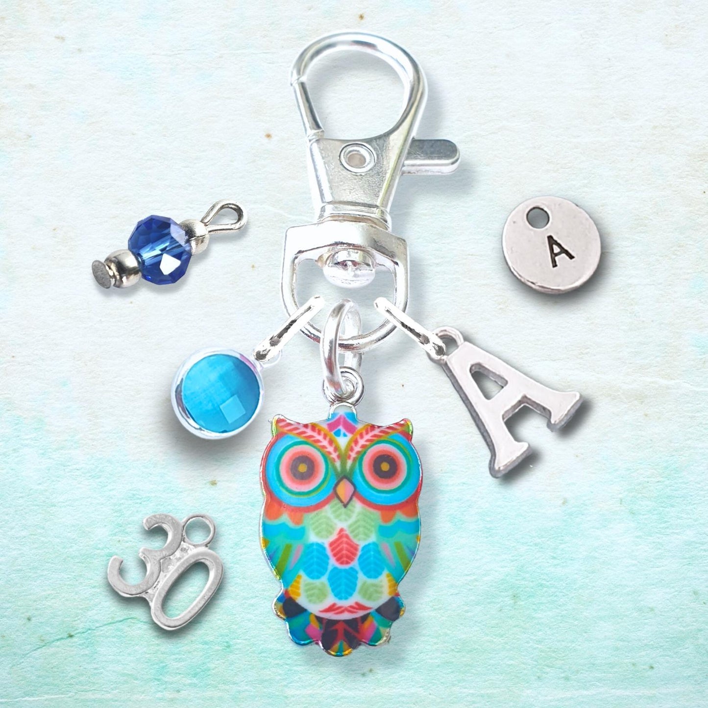 A silver-toned keychain or bag charm set displayed on a light blue textured background. The set includes a colorful owl charm, a silver letter 'A' charm, a silver '30' charm, a blue faceted bead charm, and a silver initial 'A' disc charm.