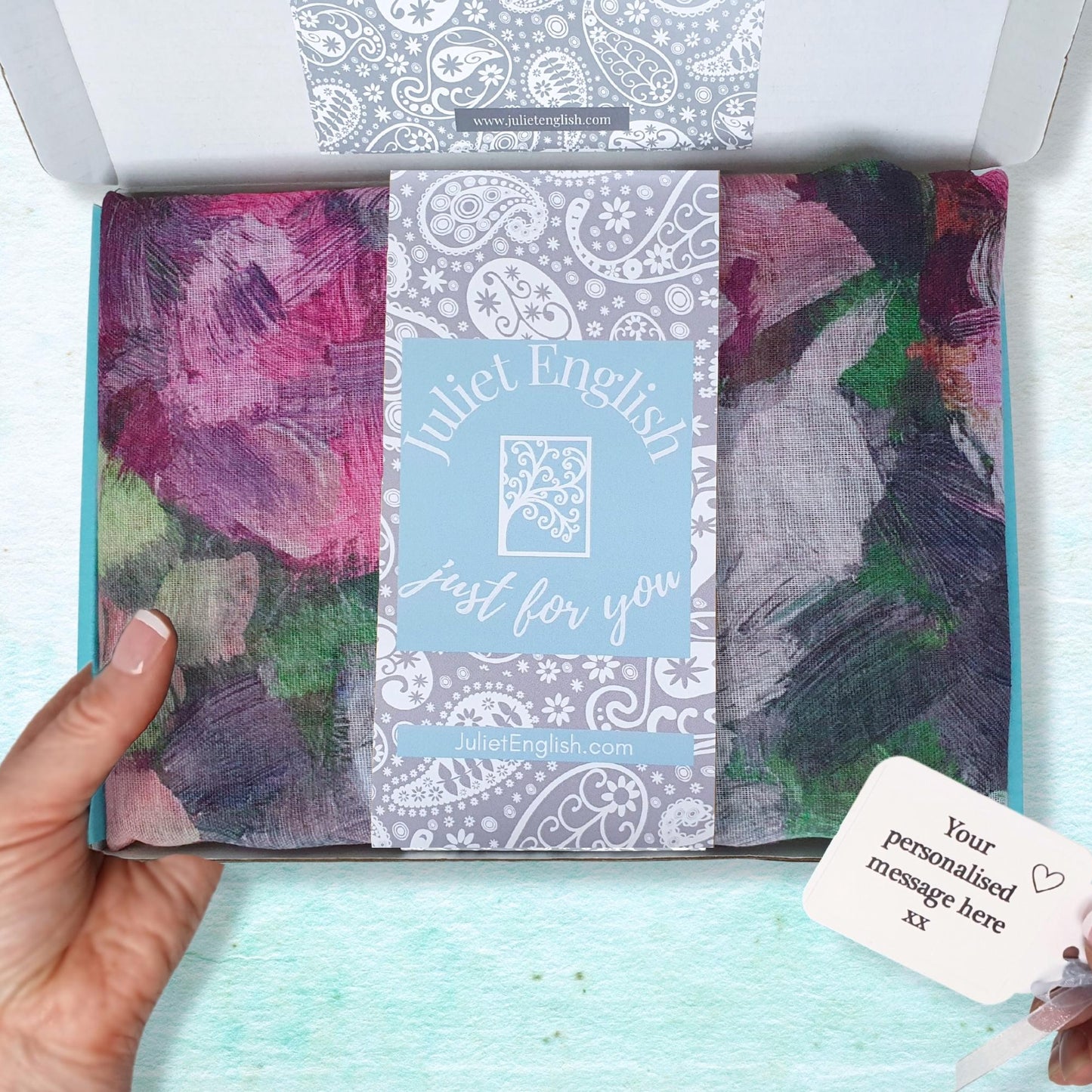 Hand holding a wrapped scarf in a gift box with a visible brand label against a light blue background