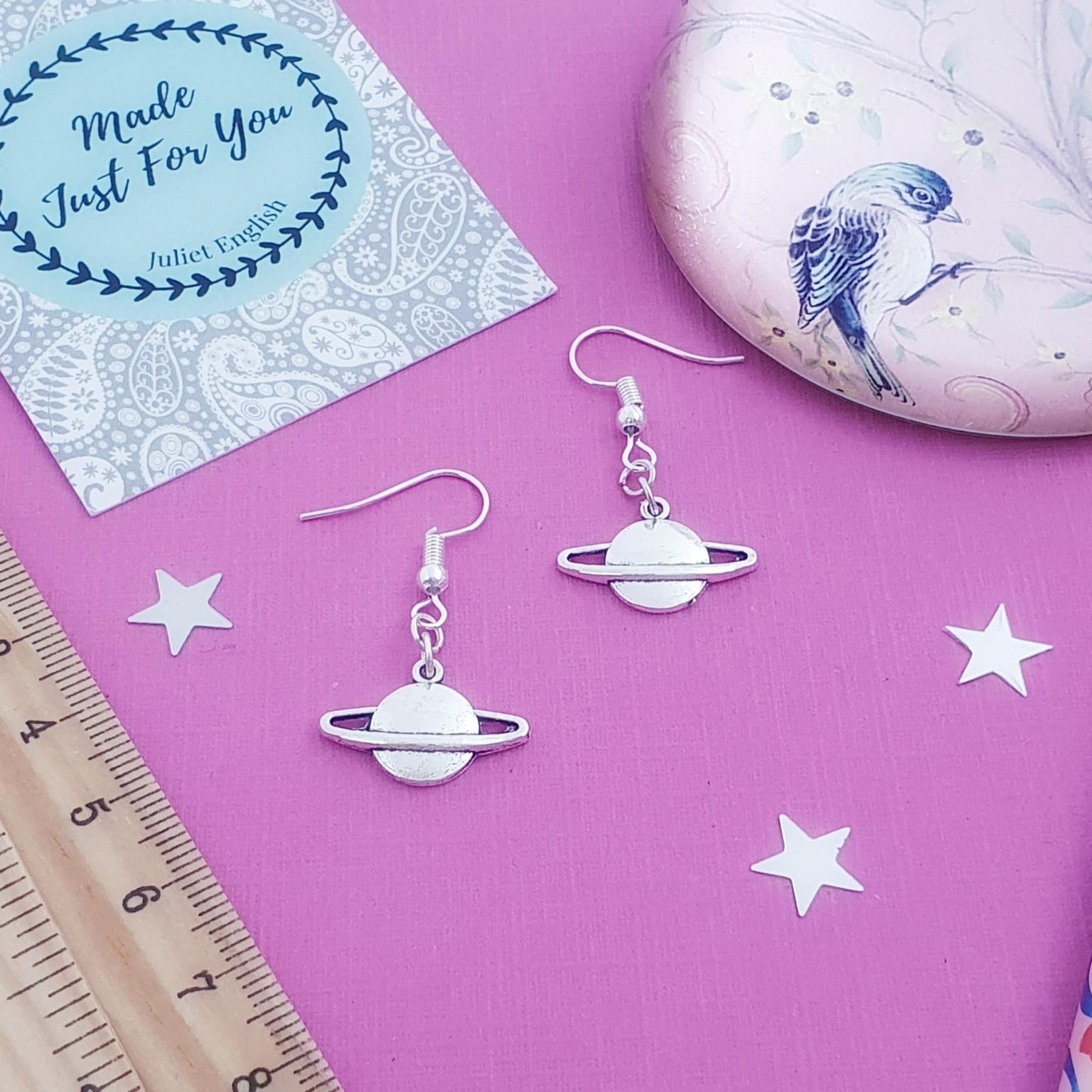 Silver planet-shaped earrings on a pink surface with a card and ruler.