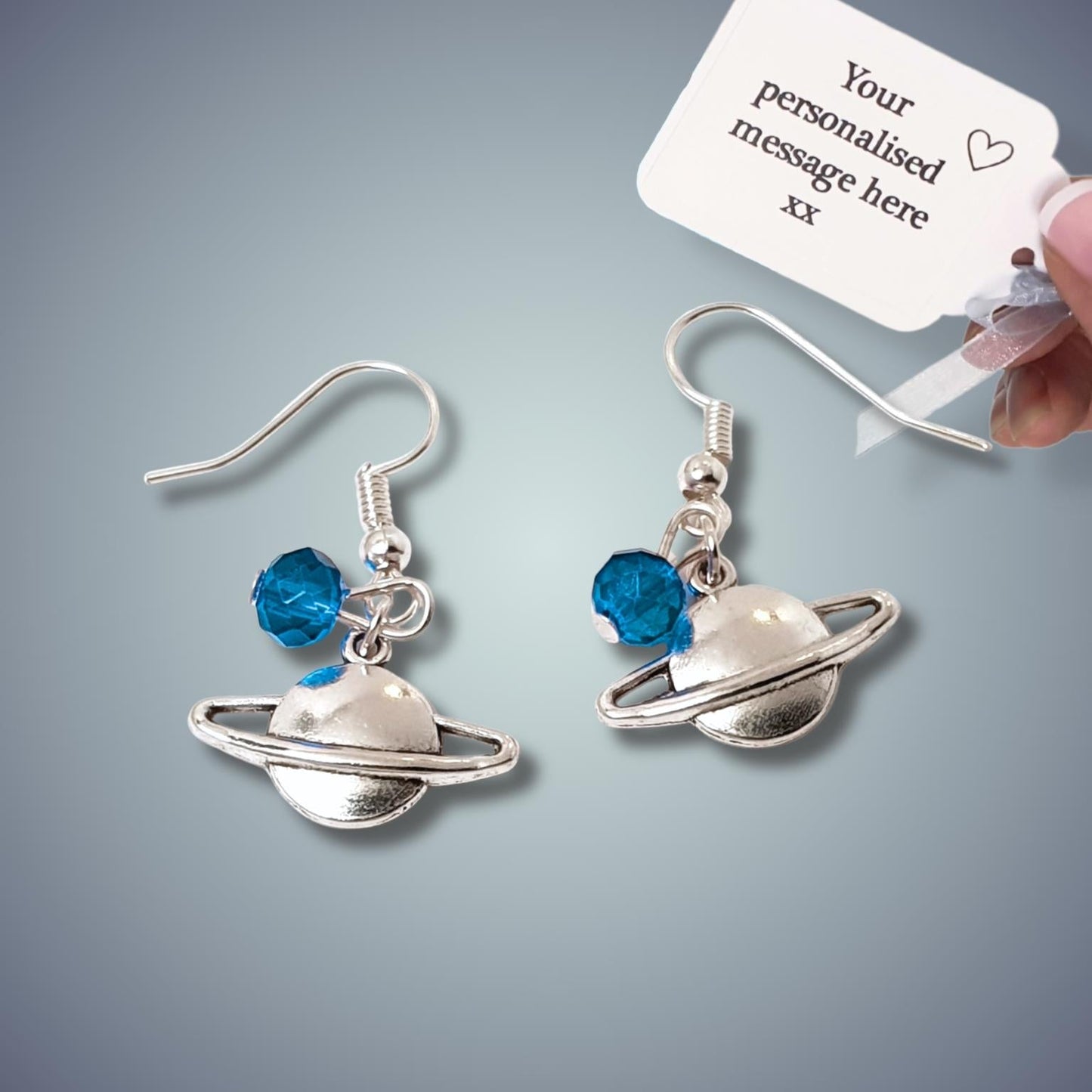 Silver Saturn-shaped earrings with blue gemstones on a gray background, held by a hand.