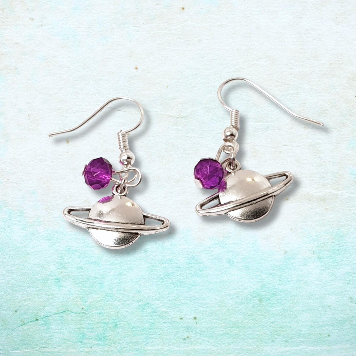Silver Saturn-shaped earrings with purple beads on a light blue and white background
