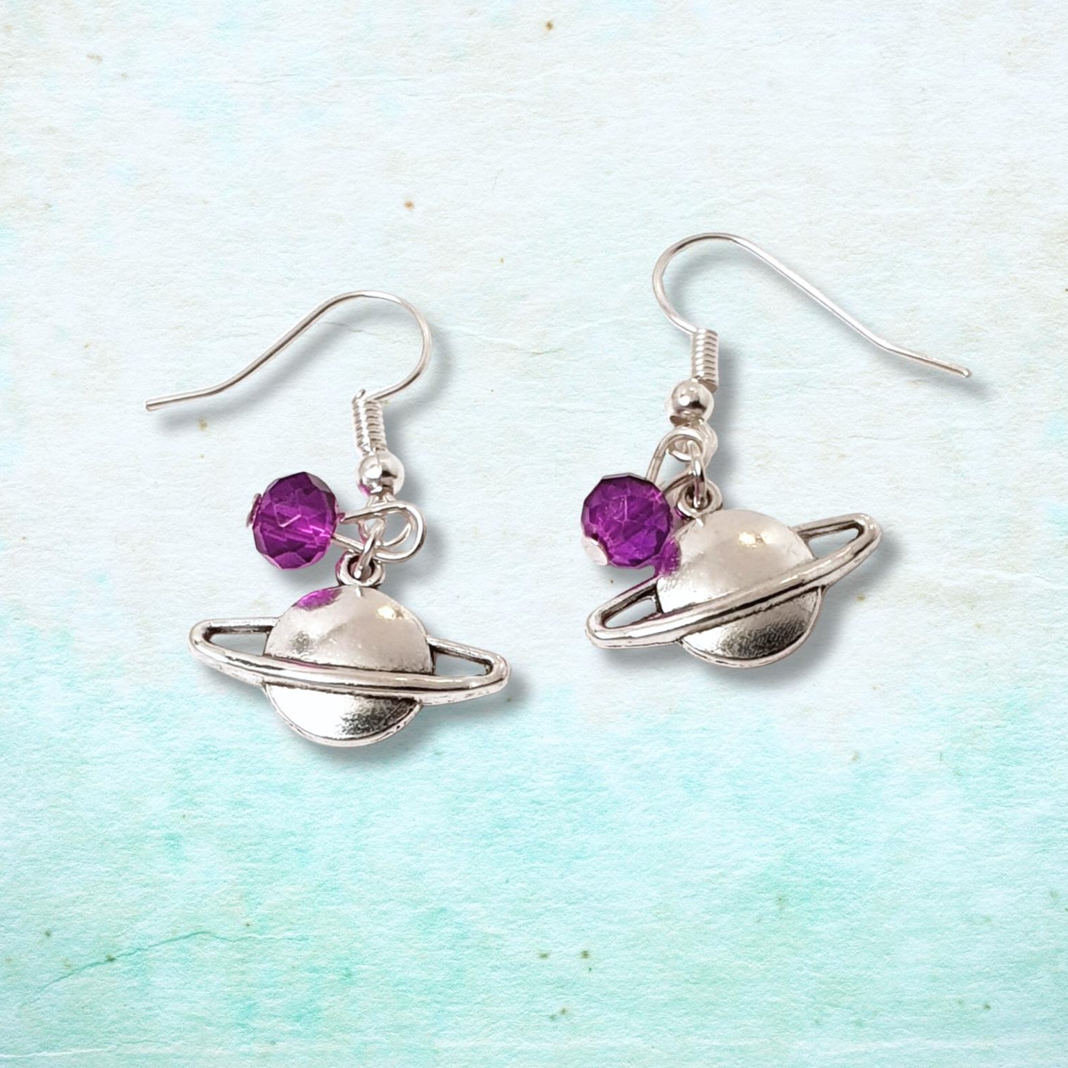 Silver Saturn-shaped earrings with purple beads on a light blue and white background