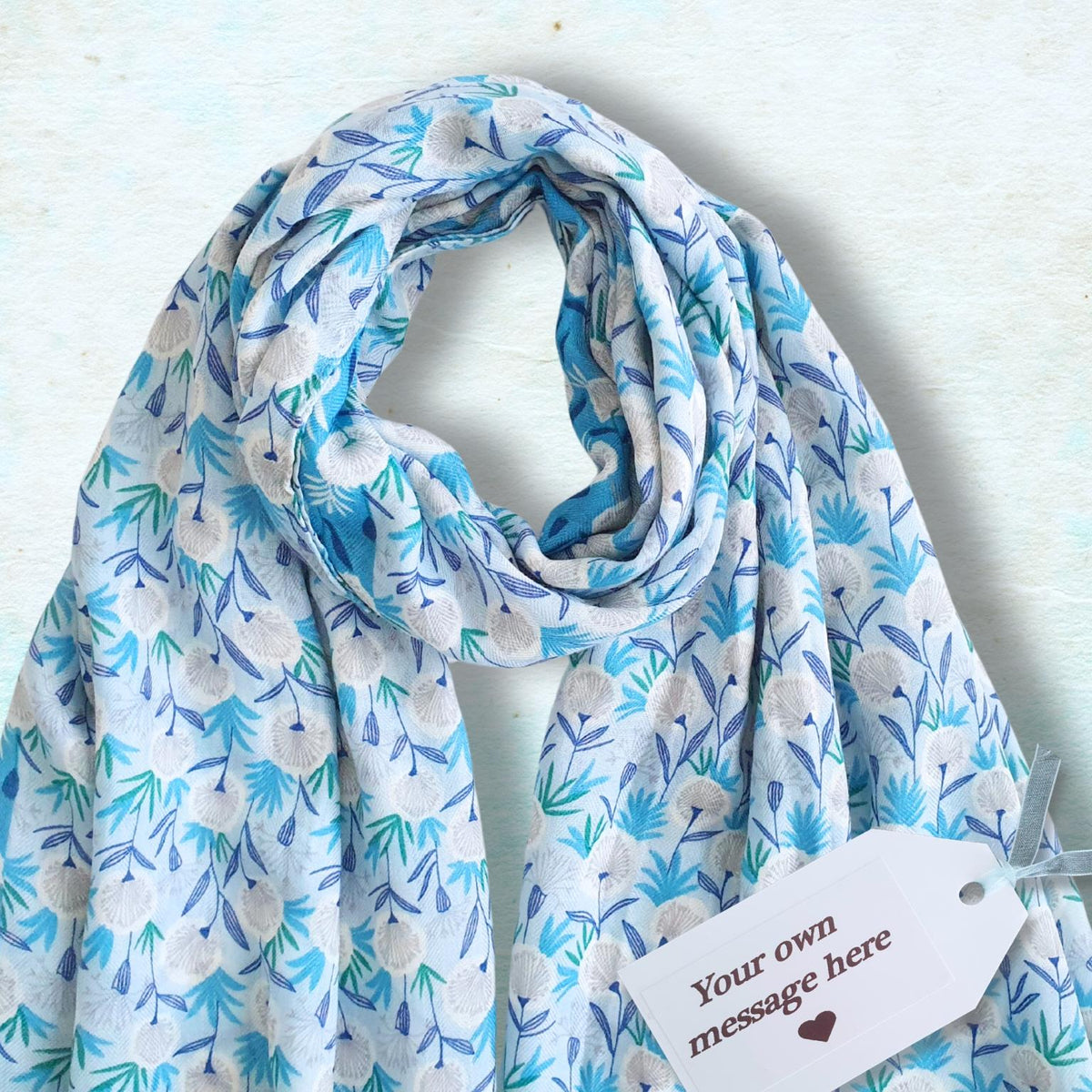 A medium weight scarf with a blue and white floral pattern, draped on a textured light blue background with a white customizable gift tag attached.