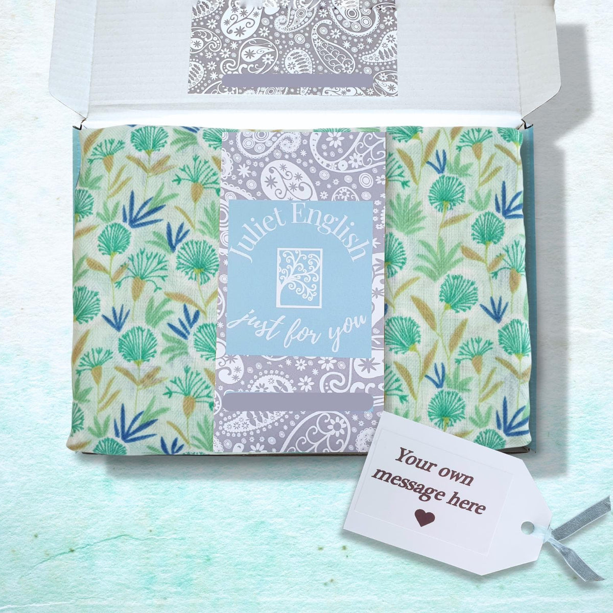 A gift box with scarf featuring green and blue botanical patterns on a light background, with a central light blue panel reading 'Juliet English Just for you' and a small white gift tag attached.