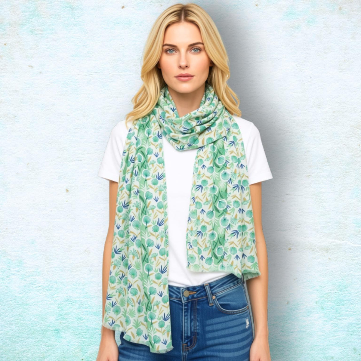 A blonde woman wearing a green and blue patterned scarf over a white t-shirt and blue jeans, standing against a textured light blue background.