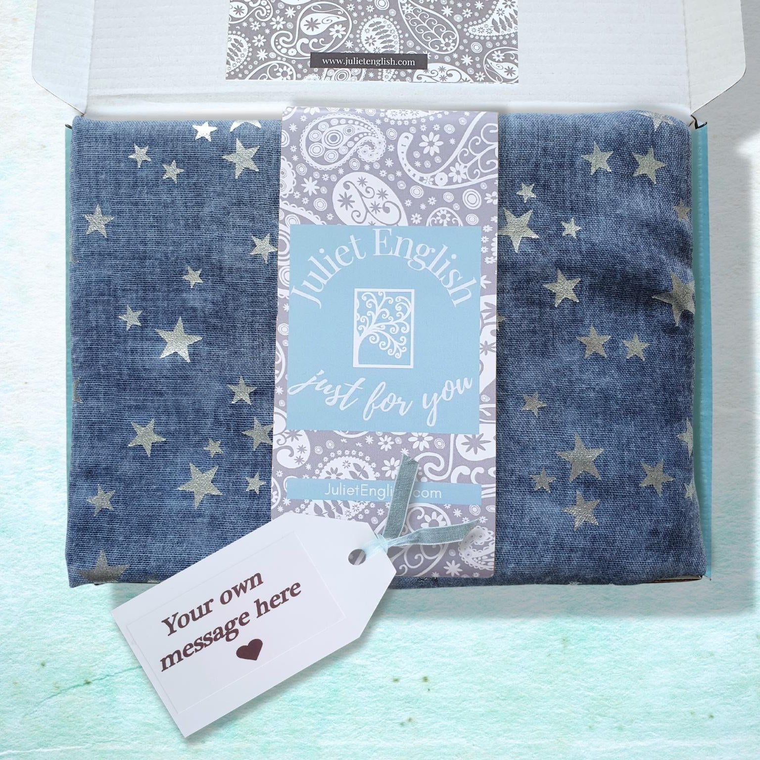A folded scarf in a blue gift box with metallic silver star patterns, wrapped in a decorative paper band that reads 'Juliet English just for you'. A white gift tag with a customizable message is attached, and the box is shown inside its open packaging on a textured light blue background.