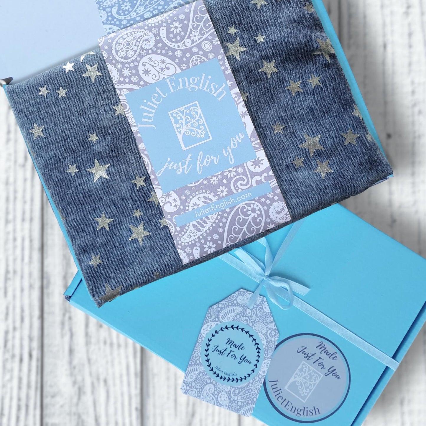 A folded blue patterned scarf with silver star prints and a central paper label reading 'Invited English Just for you,' arranged on top of a bright blue gift box tied with a ribbon and tag on a white wood background.