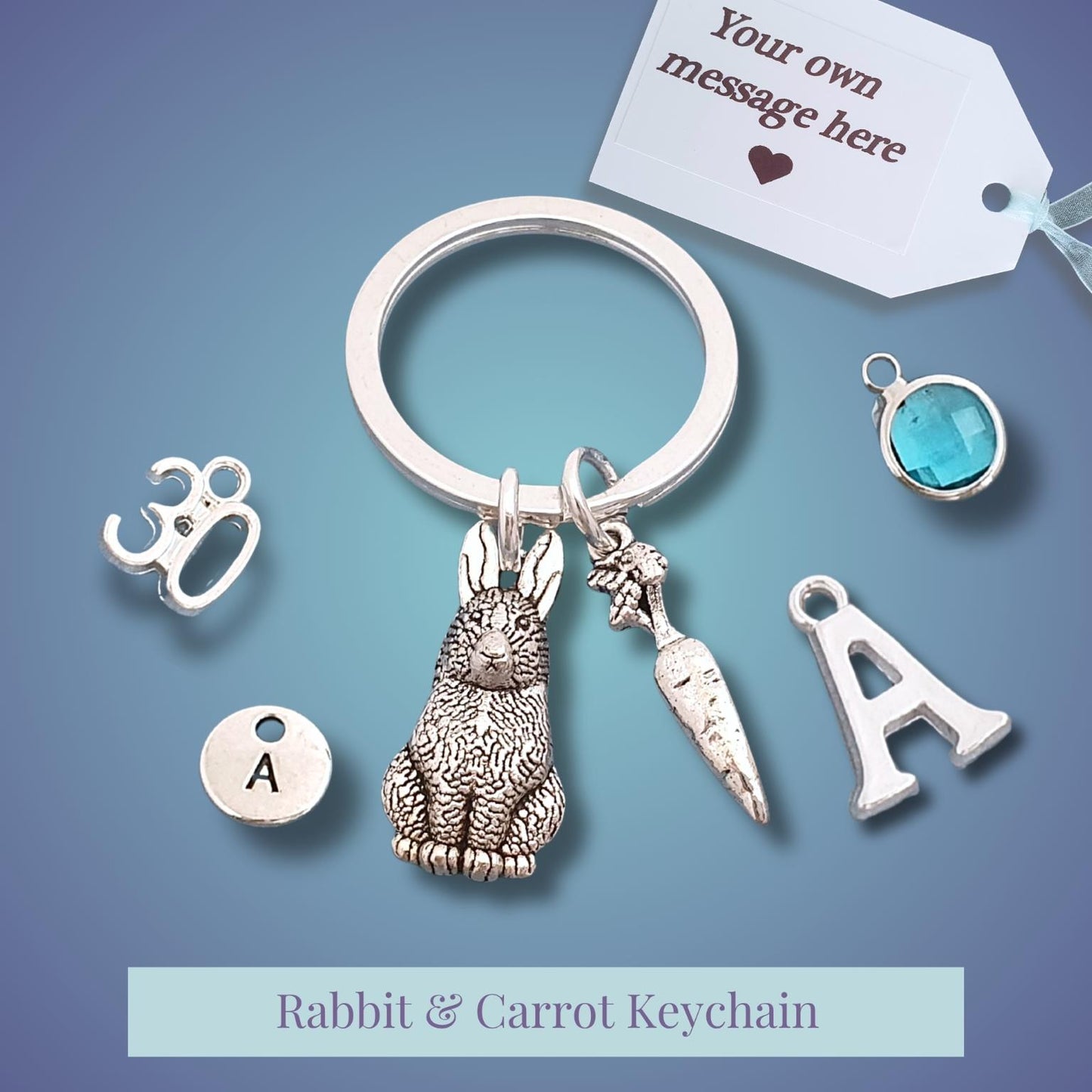 Silver rabbit and carrot keychain with dangling charms, shown on a blue gradient background with a white tag that reads 'Your own message here'.