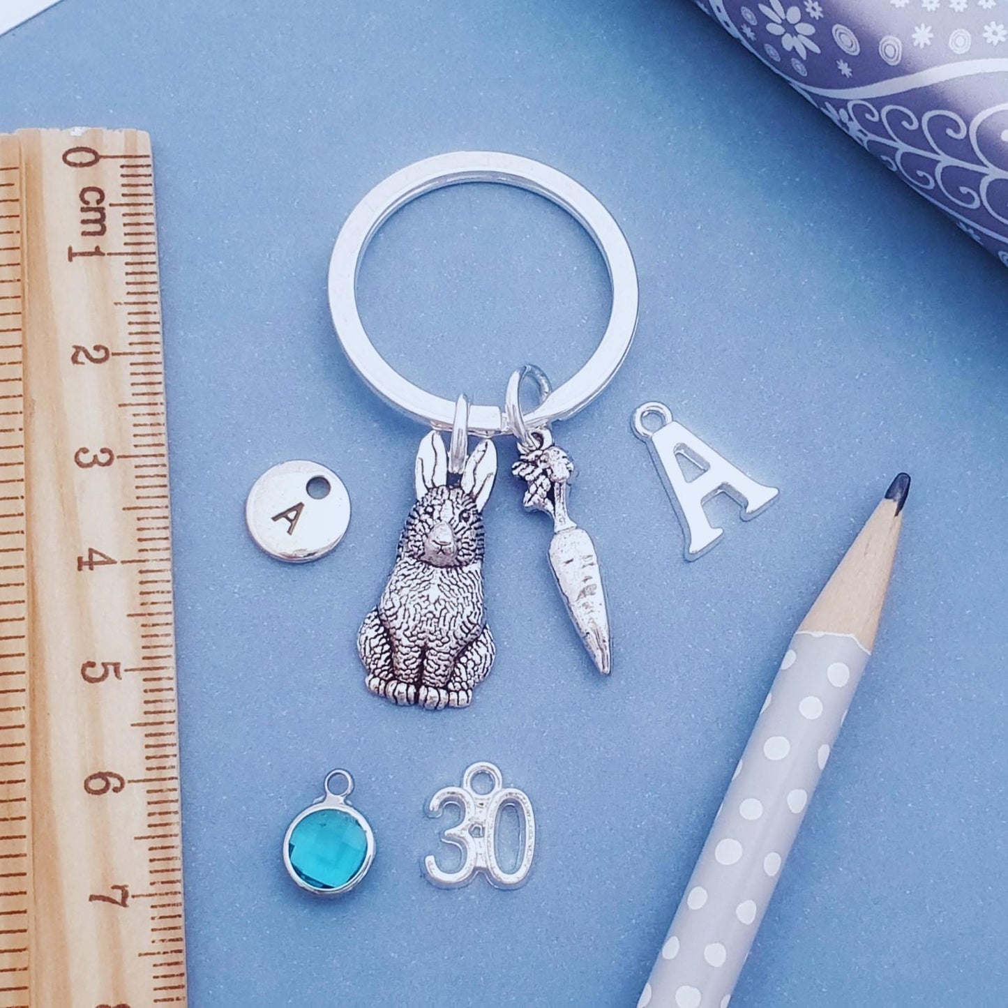 A silver keychain with charms including a rabbit, carrot, letter 'A', and '30', displayed on a blue background with a wooden ruler, pencil, and patterned ribbon.