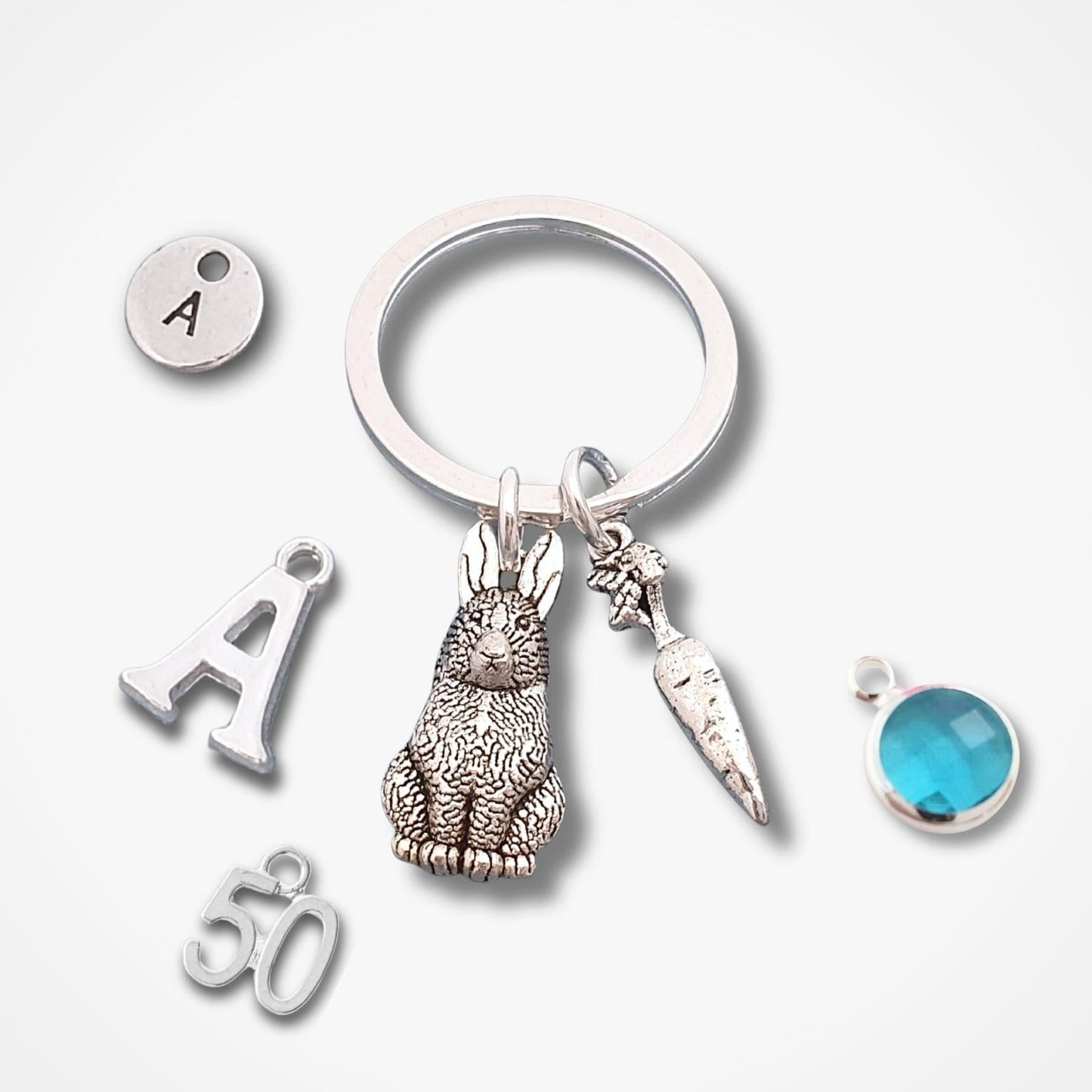 A silver keychain with multiple charms, including a detailed rabbit, a carrot, a small oval stone with the letter 'A', a letter 'A' charm, a '50' charm, and a small round blue gemstone, displayed on a clean white background.
