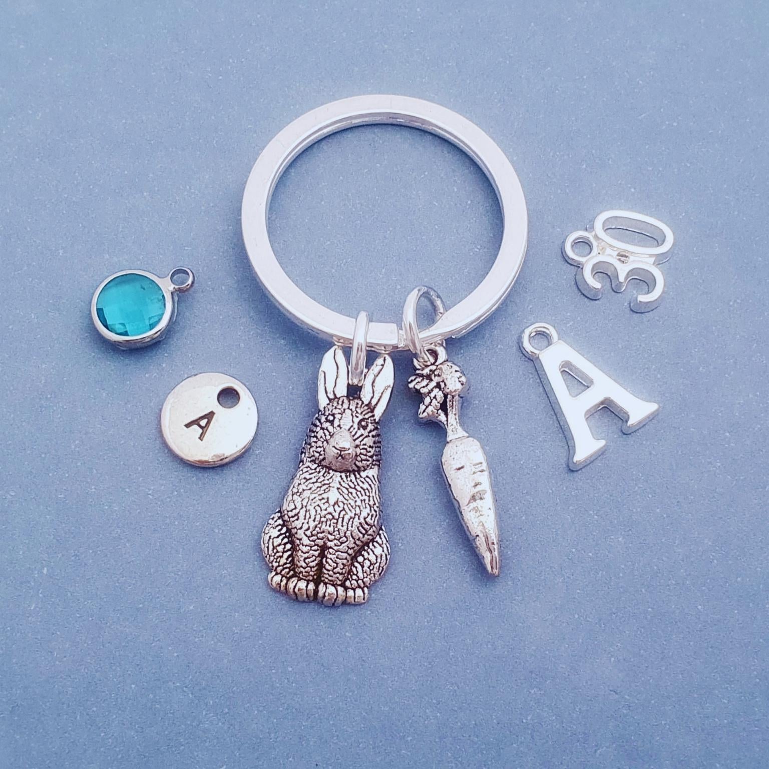 A silver keychain with a rabbit and carrot charm, displayed on a textured blue background alongside customizable charms including an initial 'A', a turquoise stone, and a '33' charm.