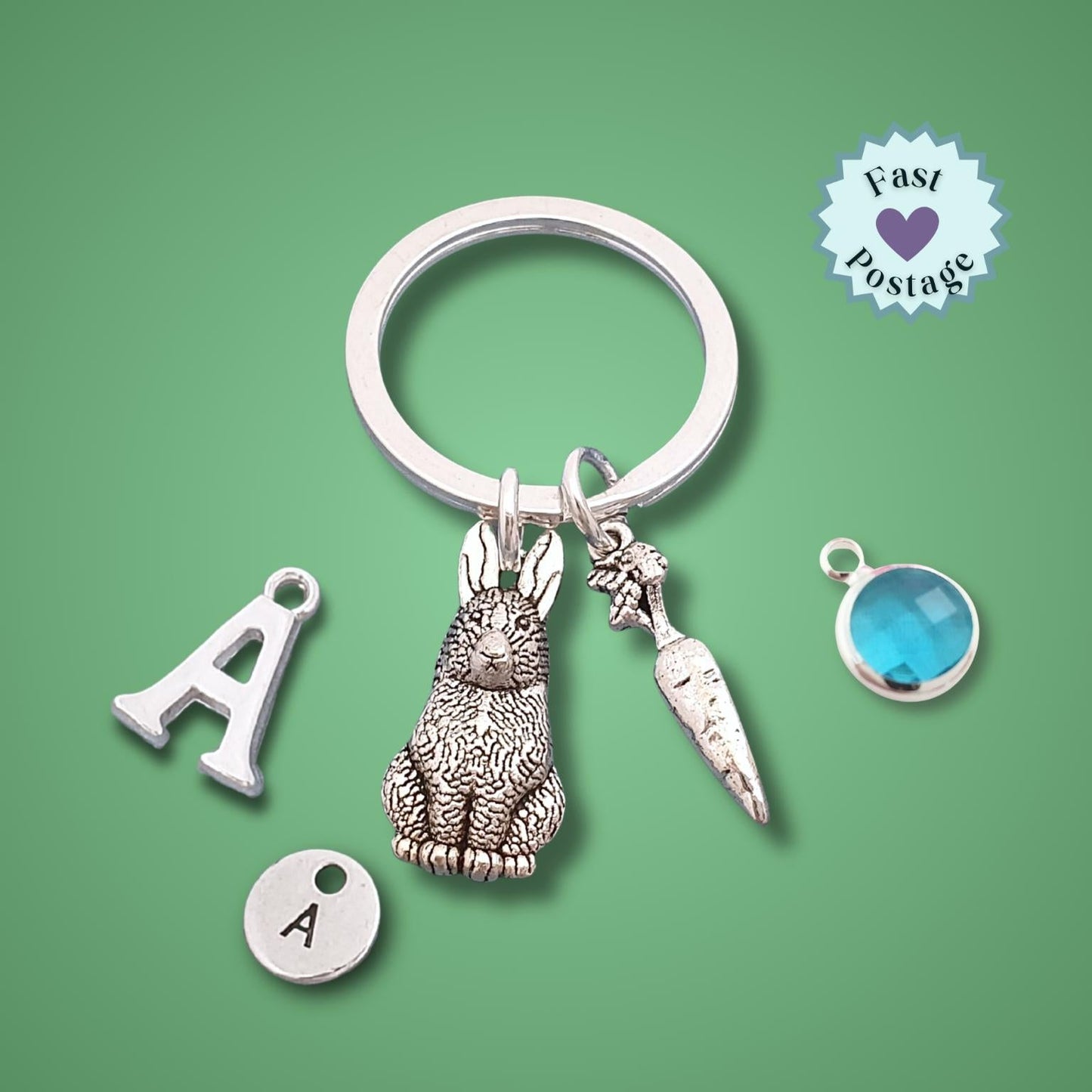 A silver-toned keychain with a rabbit charm, carrot charm, letter A charms, and a blue circular stone charm, displayed on a solid green background with a 'Fast Postage' badge.