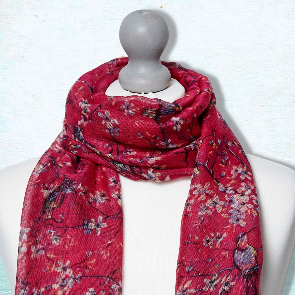 A red scarf with a floral and bird pattern draped on a white mannequin torso against a textured light blue background.
