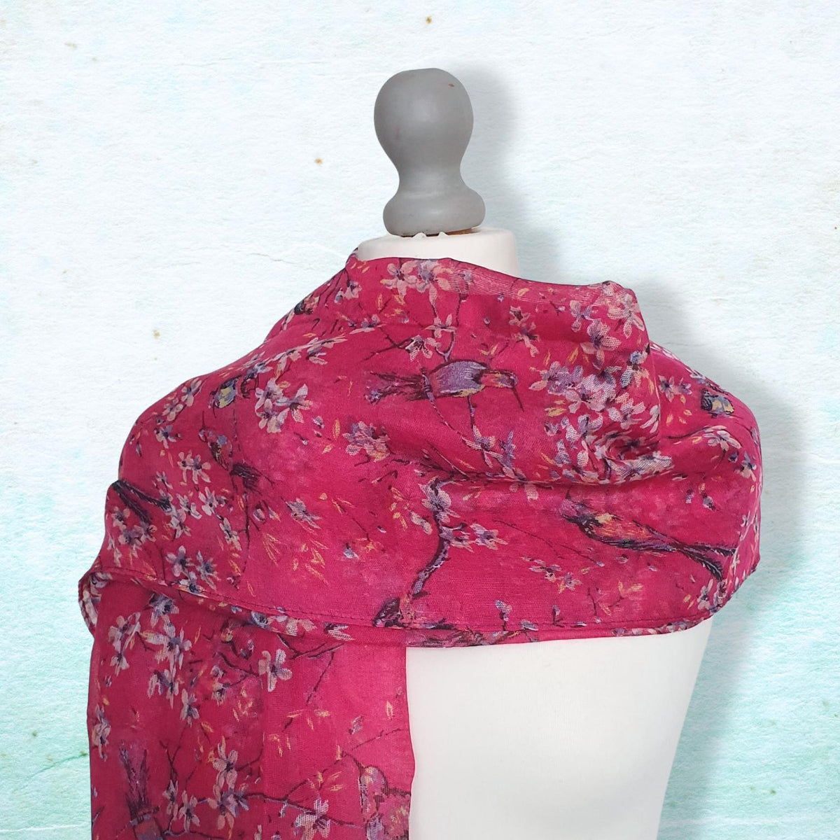 A bright pink scarf with a cherry blossom branch pattern draped on a white dress form mannequin against a textured light blue background.