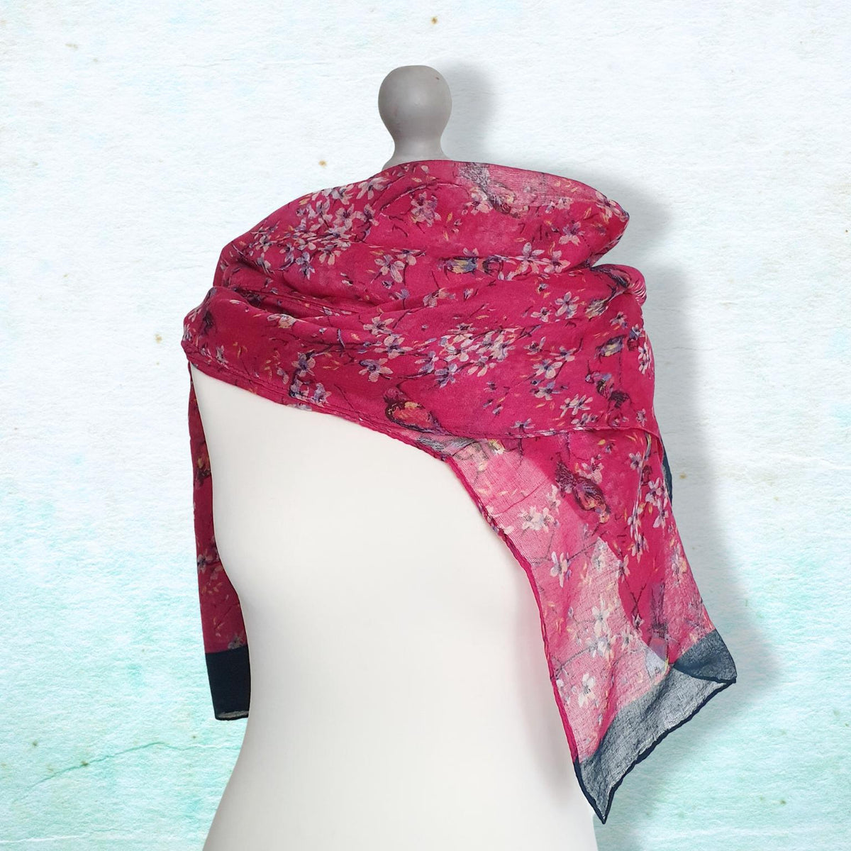 A pink floral scarf with black and white accents, draped on a white mannequin torso against a textured light blue background.