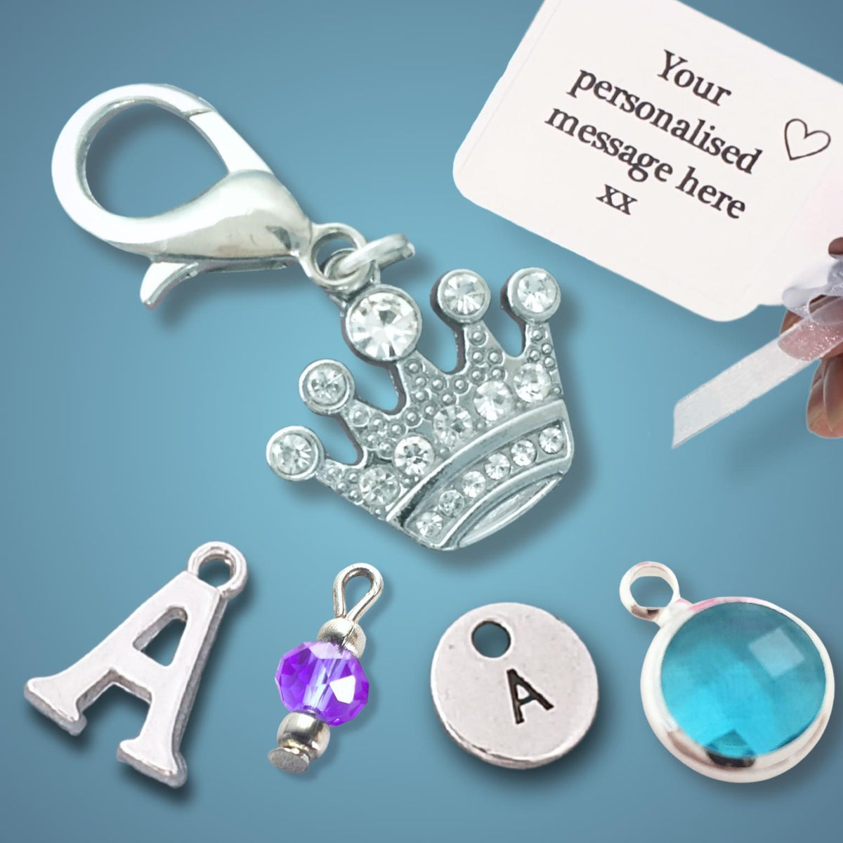 Silver initial and initial set charms for keychains, including a crown charm, letter 'A' charm, round and oval bezel-set initial 'A' charms, a purple crystal charm, and a silver lobster clasp. A hand holds a white sample tag that reads 'Your personalised message here xx'.