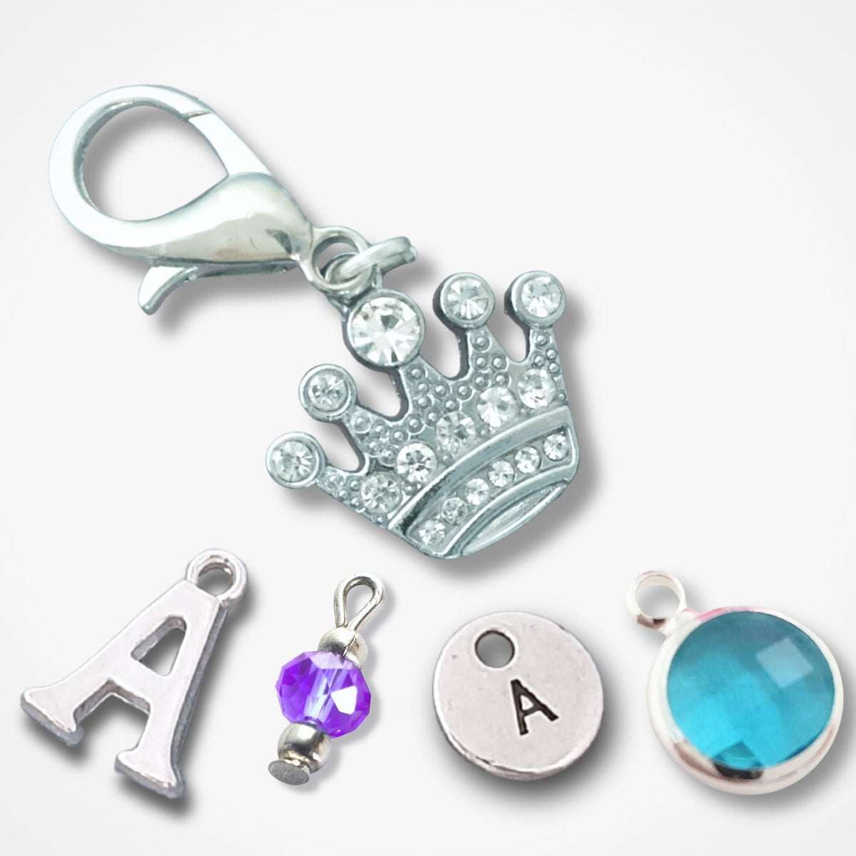 A set of silver-toned jewelry charms on a light gray background, including a crown charm with clear rhinestones, a silver letter A charm, a silver disc charm with an engraved letter A, a purple faceted crystal charm, and a round turquoise-blue faceted gemstone charm.